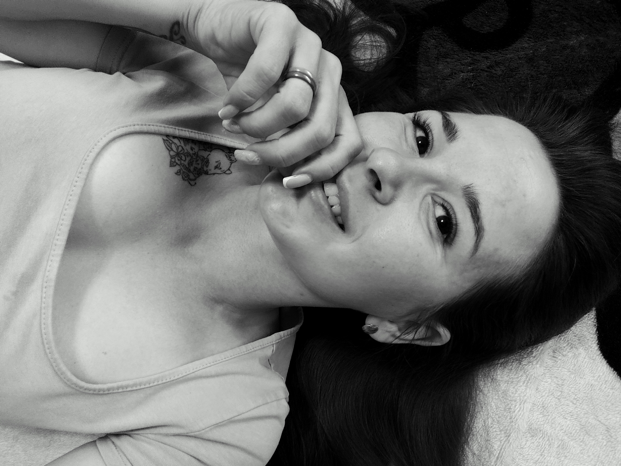 A black and white close-up image depicts a woman lying on her back, with her head tilted towards the camera. Her dark hair is spread around her head on what appears to be a textured surface. She is wearing a light-colored tank top, and a tattoo is visible on her chest. She has her hand near her face, displaying a ring on her finger and manicured nails. Her expression is pleasant, with a slight smile. The background is blurred and indistinct. There is no indication of a location in Фоки, Russia.