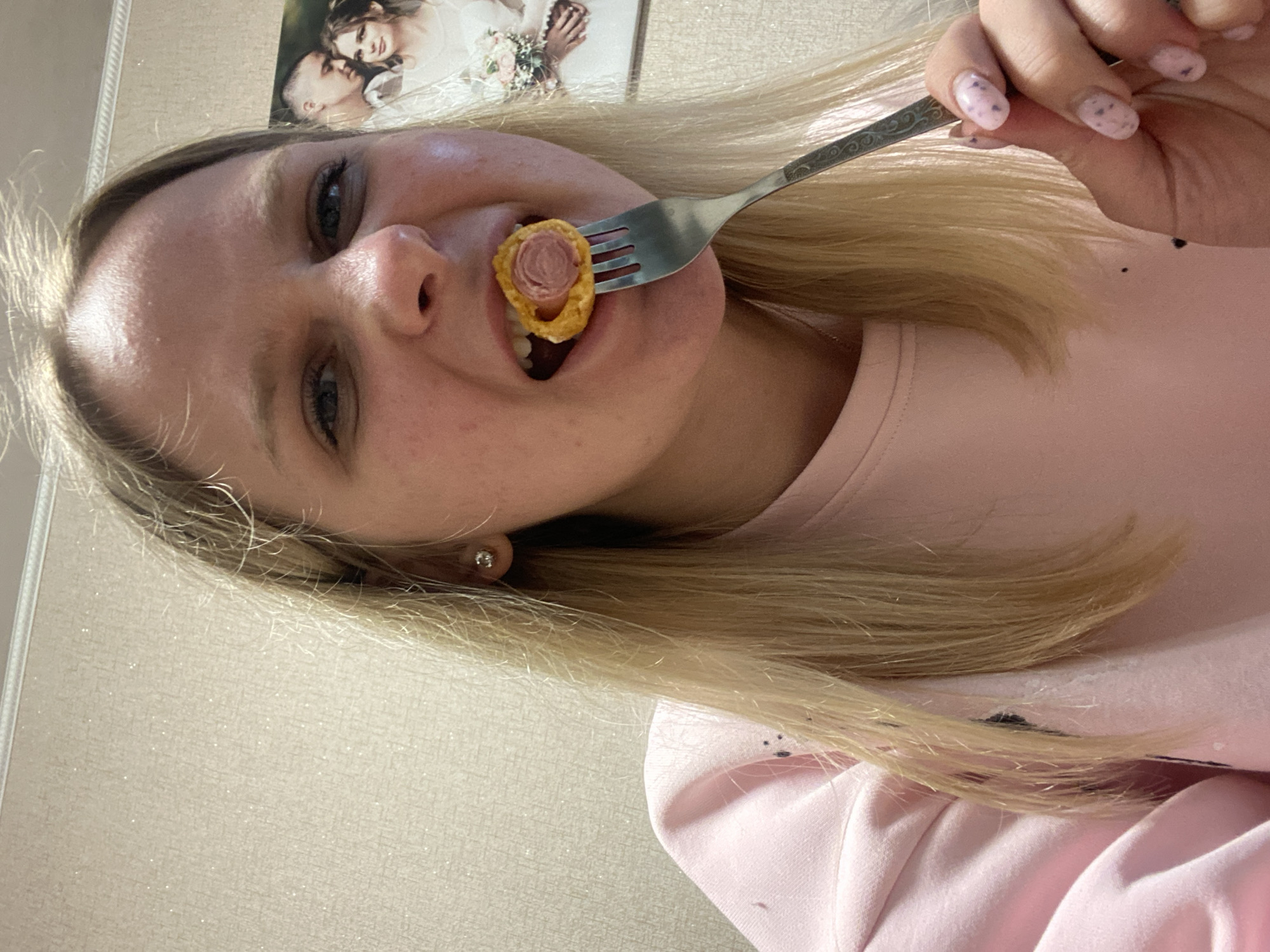 The image shows a young woman with long blonde hair and fair skin, captured in an indoor setting in Chaikovsky, Russia. She is looking directly at the camera with a slight smile. She holds a metal fork with a decorative handle to her mouth, on which is a small piece of food resembling a mini sausage or hot dog wrapped in golden-brown dough. She appears to be in the process of taking a bite.
She is wearing a light pink long-sleeved top. Her fingernails are painted light pink with small dark speckles, and she has a small silver stud earring in her left ear. The background features a light-colored, textured wall. Above her head, a framed photograph is visible on the wall, depicting a man with a shaved head and a woman with dark hair embracing, suggesting a wedding portrait. There is no visible text in the image. The scene is brightly lit, consistent with an indoor environment.