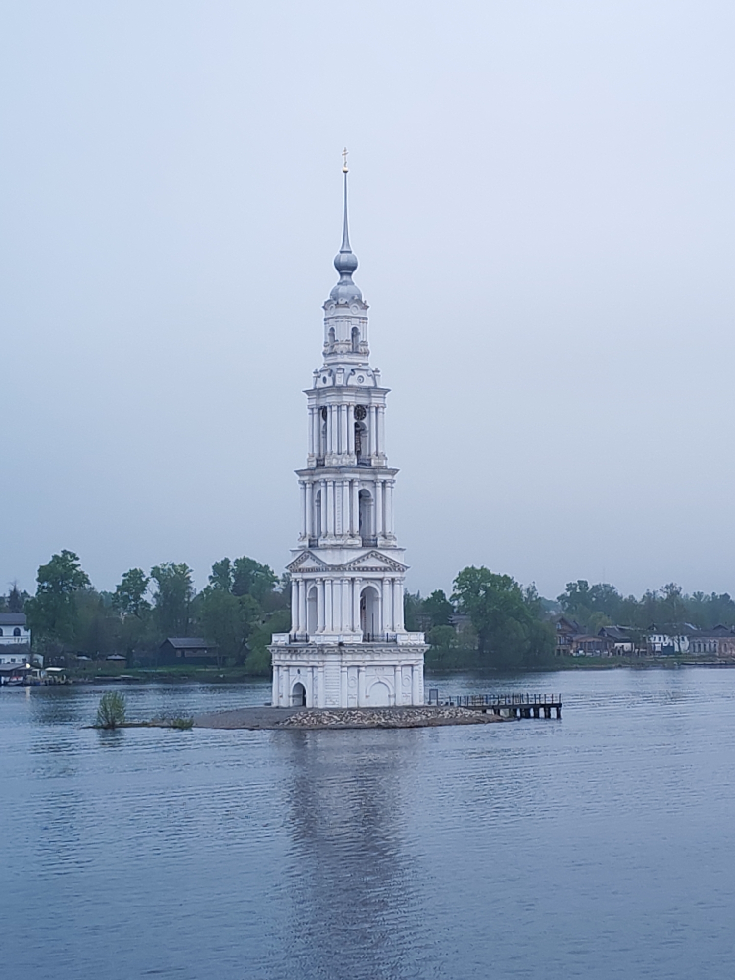 The image shows the Kalyazin Bell Tower in Kalyazin, Russia. The white bell tower is a prominent landmark, standing in the middle of a body of water. The water surrounds the tower and reflects its image. A small area of land is visible around the base of the tower. A dock extends into the water near the tower. In the background, buildings and trees line the shore. The sky is overcast, with a muted color palette throughout the scene.