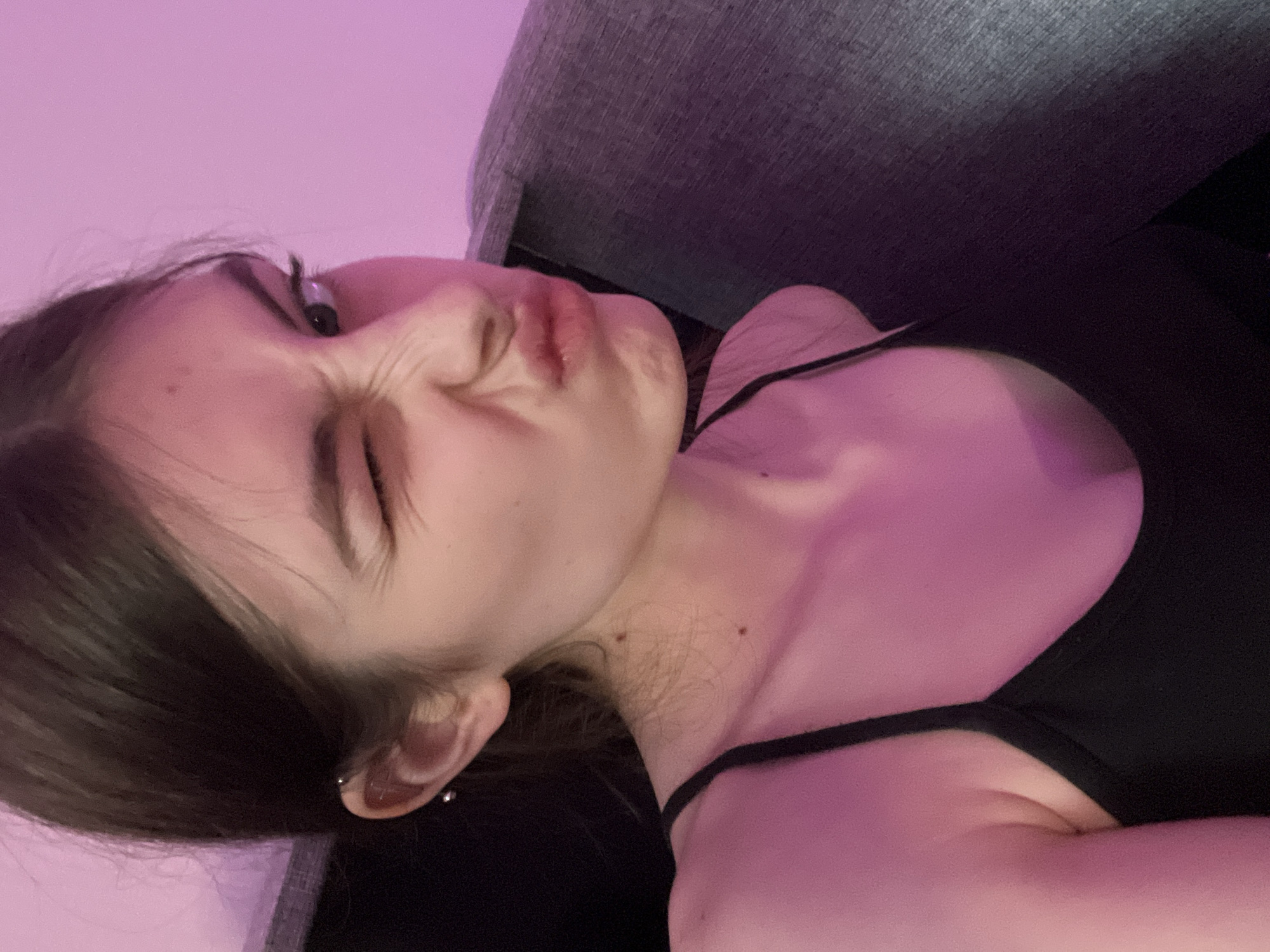 A young woman with dark hair is lying down, possibly on a couch, with a pink light illuminating the scene. Her eyes are closed, and her face is contorted into a grimace. She is wearing a black top. An ear piercing is visible in her left ear. The setting is indoors, with a gray surface behind her. Location: Unknown, Russia.
