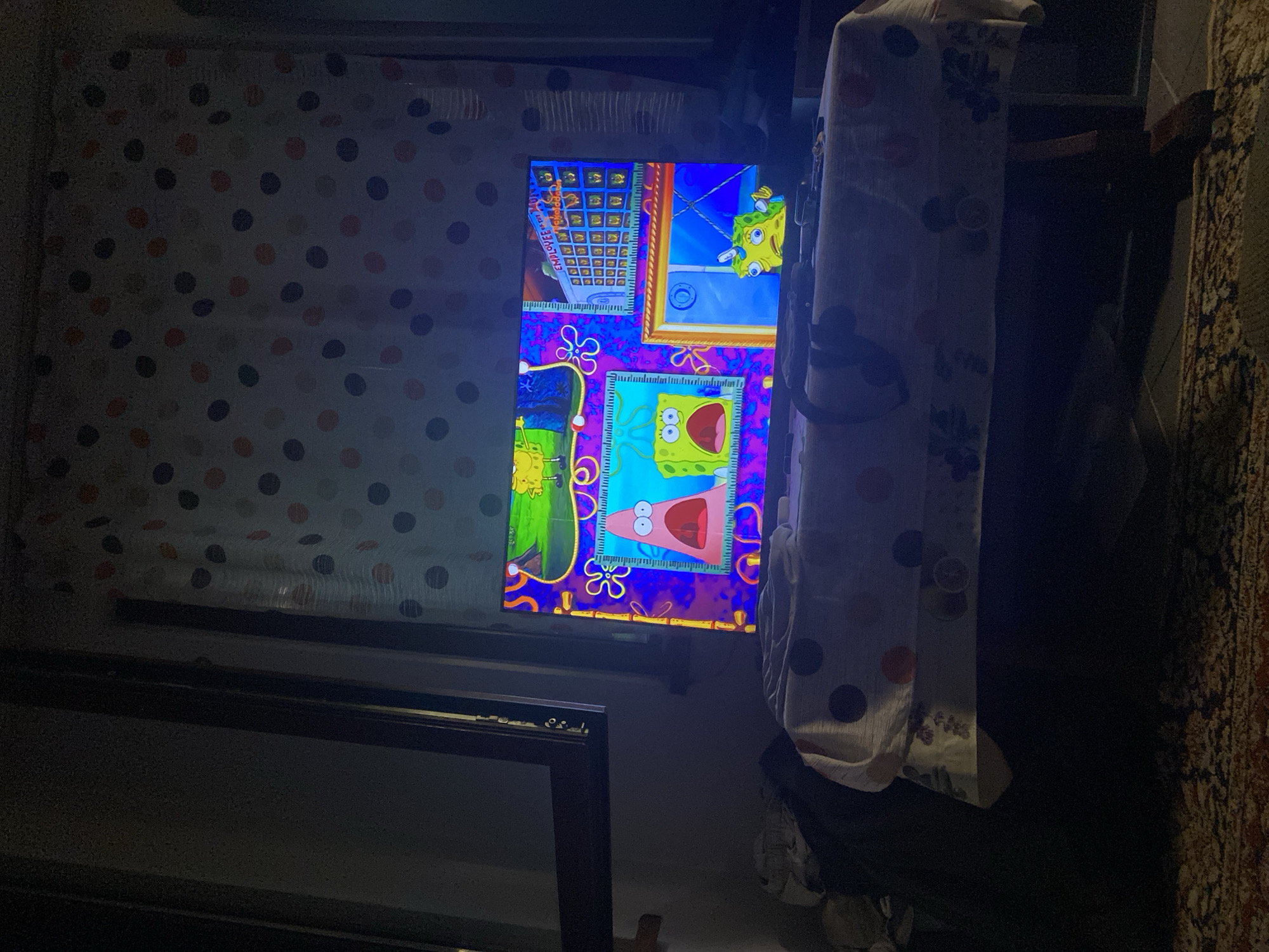 A television screen displays a scene from the cartoon SpongeBob SquarePants. The screen is illuminated, casting a colorful glow on the surrounding dimly lit room. The television is positioned on a table covered with a patterned tablecloth. A dark curtain with polka dots hangs in the background. The scene on the TV features various characters from the show in a vibrant, animated setting.