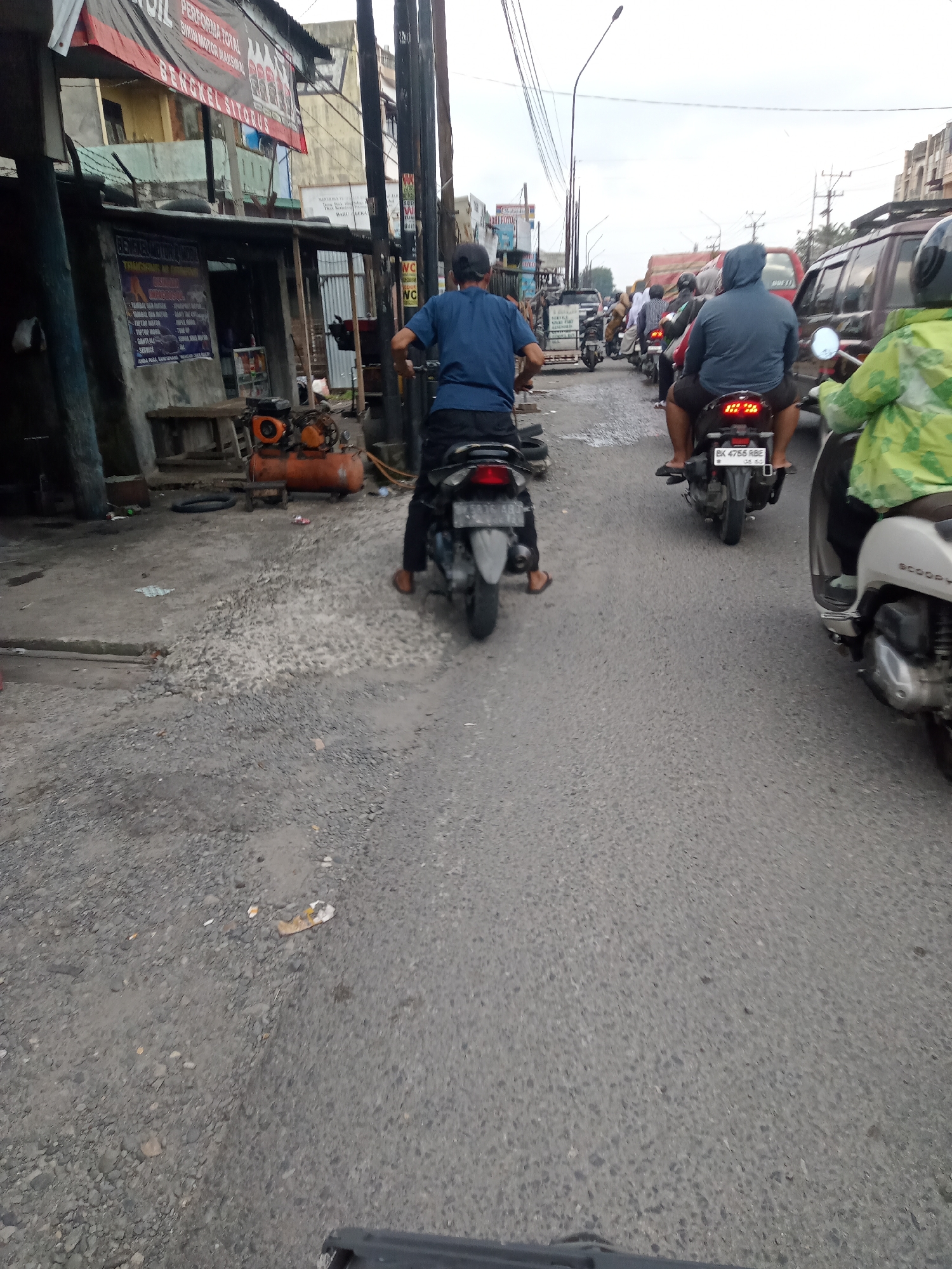 A motorcycle is parked on a street in Sei Semayang, Indonesia, with other motorcycles and vehicles in the background. The motorcycle is black and has a license plate that reads "BK 4765 RBE." It is parked on the left side of the road, and there is a man wearing a blue shirt and a black hat sitting on it. The man is looking forward. In the background, there are other motorcycles and cars, as well as a building with a red sign. The road is paved with asphalt, and there is a pile of gravel on the left side of the road. The sky is overcast.