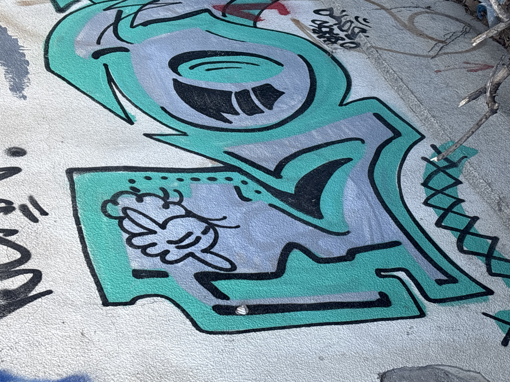The media file shows a close-up view of graffiti on a textured concrete surface. The graffiti features bold outlines and fills in shades of teal, light purple, and black, creating a stylized character or abstract design. In the lower portion of the image, a small, simplified drawing of a cloud with a face, possibly a character, is visible within a teal-bordered area. The graffiti itself is vibrant and appears to be relatively new, with no significant signs of weathering.
To the right of the main graffiti, a dry, twig-like branch rests on the surface, obscuring a small portion of the artwork and some faint, less defined markings on the concrete. These markings appear to be other, older graffiti or perhaps scuffs. The overall setting seems to be outdoors, indicated by the presence of the branch and the textured concrete, which could be a wall or pavement. There is no discernible text within the graffiti itself. The lighting suggests it is daytime, with no strong shadows or indications of specific weather conditions. The image is a still shot, capturing a static scene.