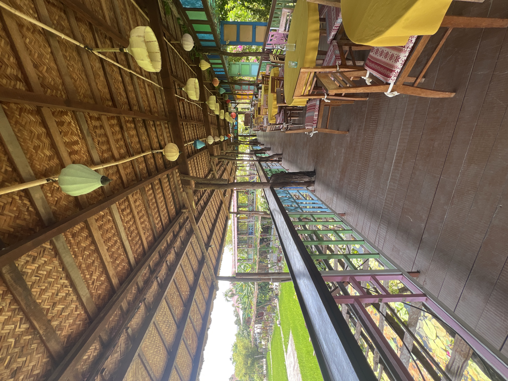 This image depicts a covered outdoor dining area with a view of lush greenery and what appears to be a rice paddy field, characteristic of the rural outskirts of Hoi An, Vietnam. The structure features a natural, woven bamboo ceiling supported by wooden posts. Hanging from the ceiling are various colorful paper lanterns, adding a decorative touch. The walkway, made of wooden planks, is lined with a railing painted in pastel colors of pink, light blue, green, and yellow.
The walkway overlooks a body of water filled with lily pads, with more greenery and trees visible in the background. Further in the distance, a building with a red roof and colorful window frames can be seen.
The dining area itself is furnished with several round tables covered in bright yellow tablecloths. Each table is surrounded by wooden chairs, many of which have decorative patterned cushions. The arrangement suggests a restaurant or cafe setting. The scene is bathed in natural daylight, indicating it is daytime. There are no people visible in the image, giving the impression of an unoccupied space. The overall atmosphere is serene and suggests a tranquil dining experience.