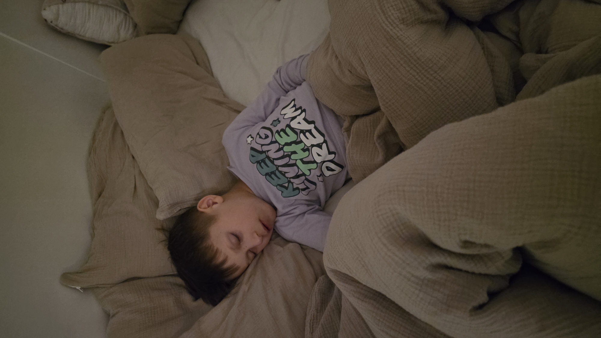 A child, wearing a lavender long-sleeved shirt with the text "KEEP DREAMING" in colorful lettering, is sleeping on a bed. The bed is covered with a beige blanket, and there are pillows in the background. The location of the image is not provided.
