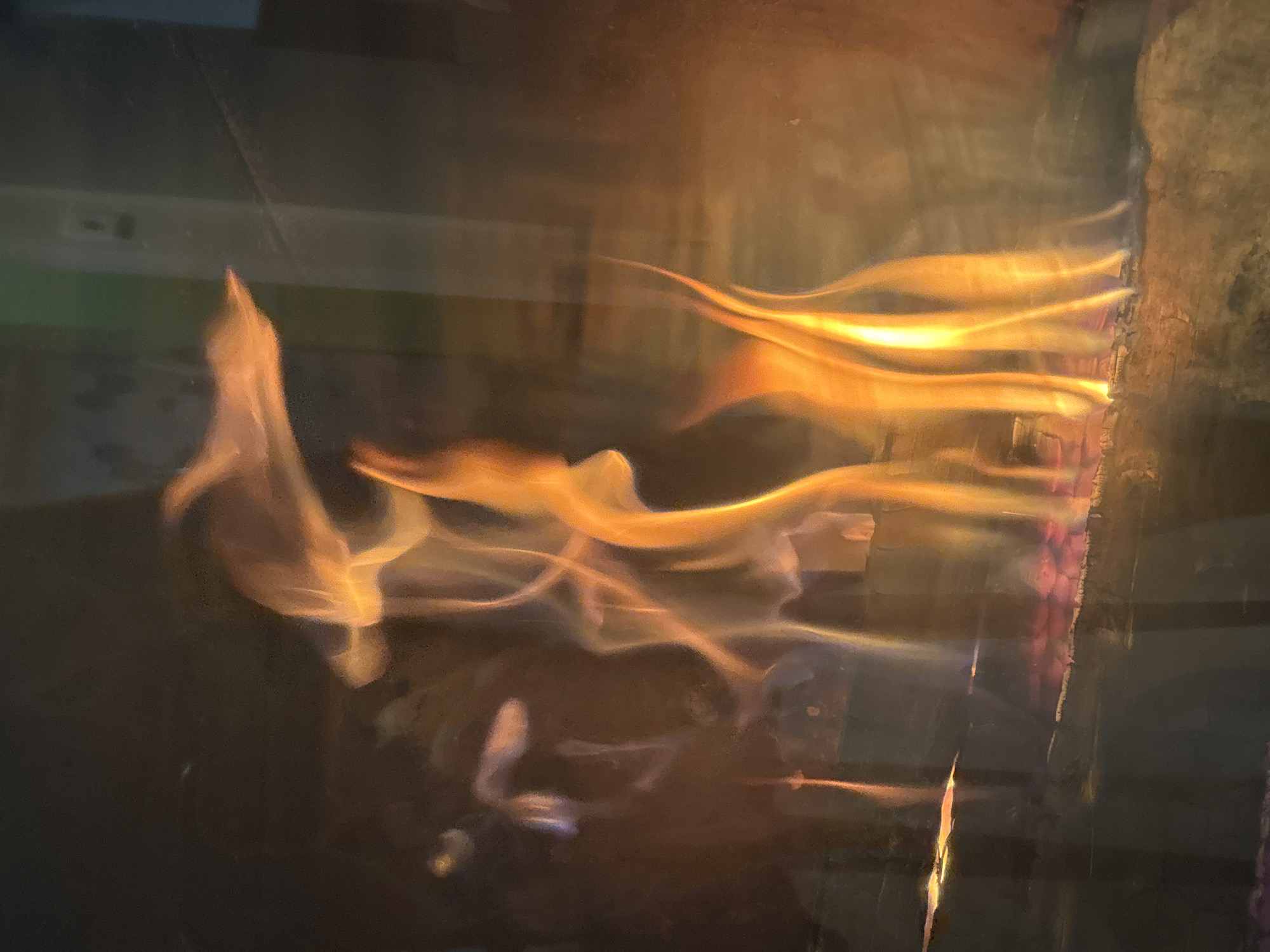 A close-up, eye-level shot captures the mesmerizing dance of flames in a fireplace. The fire is burning on logs, with bright orange and yellow hues dominating the scene. Wisps of smoke curl upwards, creating an ethereal effect. The background is dark and blurred, focusing all attention on the dynamic movement of the fire. The image appears to be taken indoors, possibly in a residential setting. No specific location in Zvenigorod, Russia, can be identified from the image.