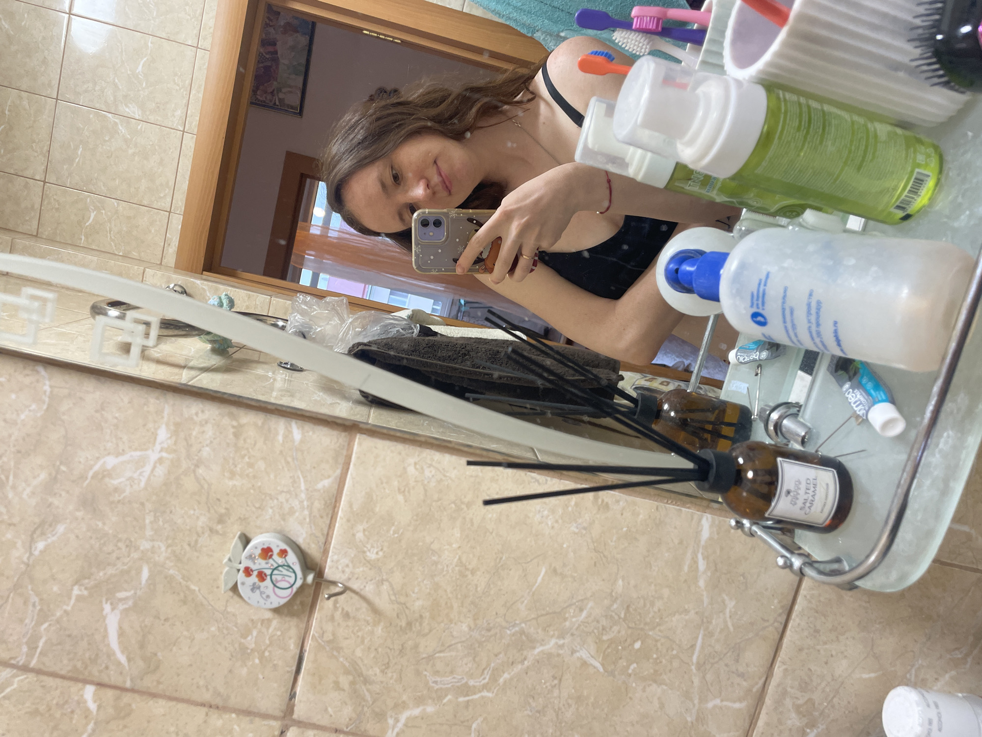 A young woman is taking a selfie in a bathroom. The woman is holding a smartphone with a decorative case. She's wearing a black top and a delicate necklace. Her hair is brown and partially pulled back. The bathroom has beige tile walls and a vanity with toiletries, including bottles of cleanser, toothpaste, and a reed diffuser labeled "SALTED CARAMEL." A toothbrush holder with several toothbrushes is also visible. A small decorative hook with a floral design is mounted on the tiled wall. A glimpse of a door and a room beyond can be seen reflected in the mirror. The city mentioned, Берёзовка, Russia, cannot be confirmed as a location based on the visual data.