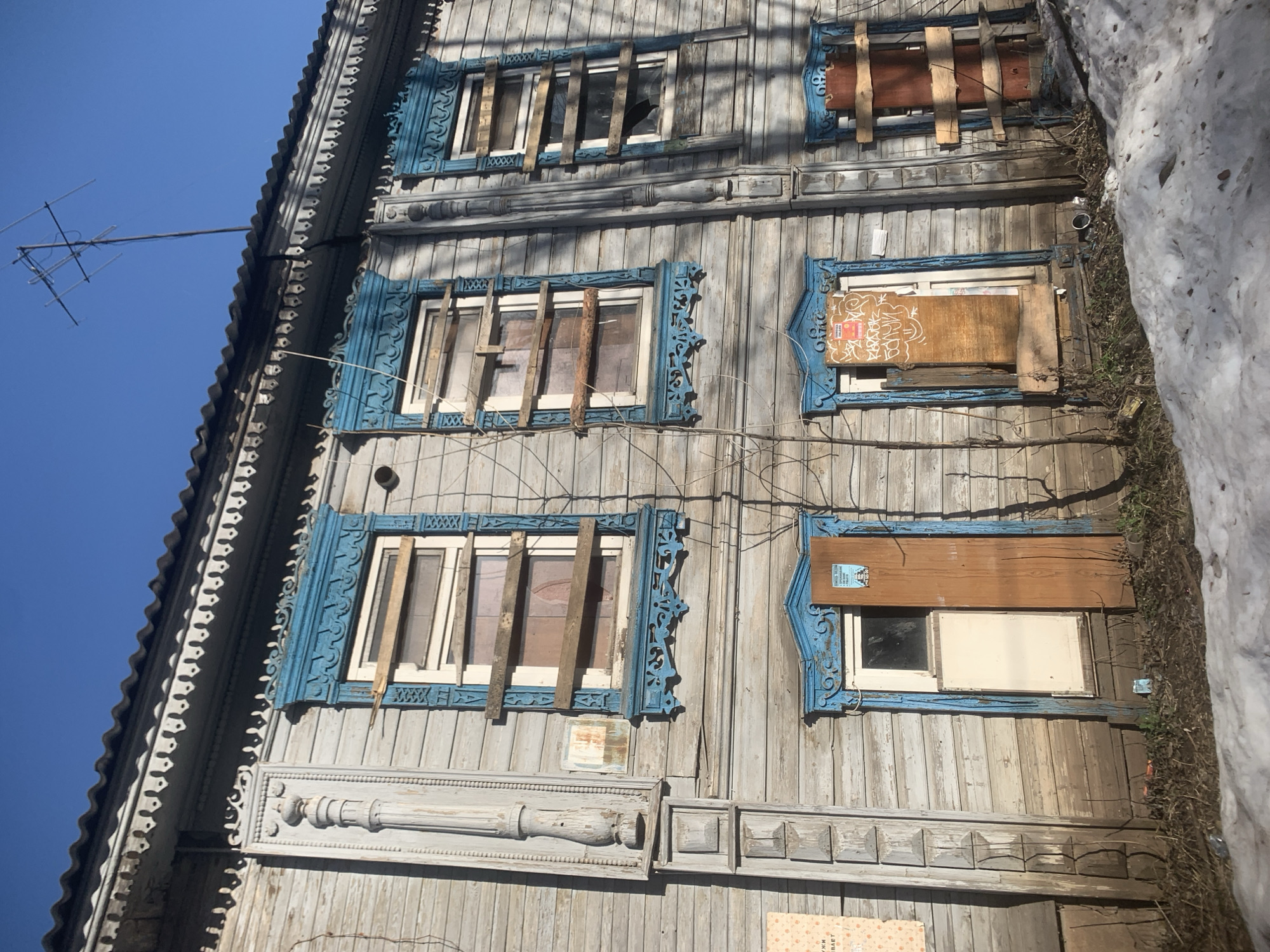 The image depicts a three-story wooden building, likely located in Izhevsk, Russia, given the architectural style. The building appears to be in a state of disrepair. The exterior is weathered, and many of the windows are boarded up or have broken panes covered with makeshift wooden planks. The window frames are painted blue, with intricate, decorative carvings. An old television antenna is visible on the roof. There is snow present on the right side of the building, suggesting a winter or early spring setting. The sky is blue.