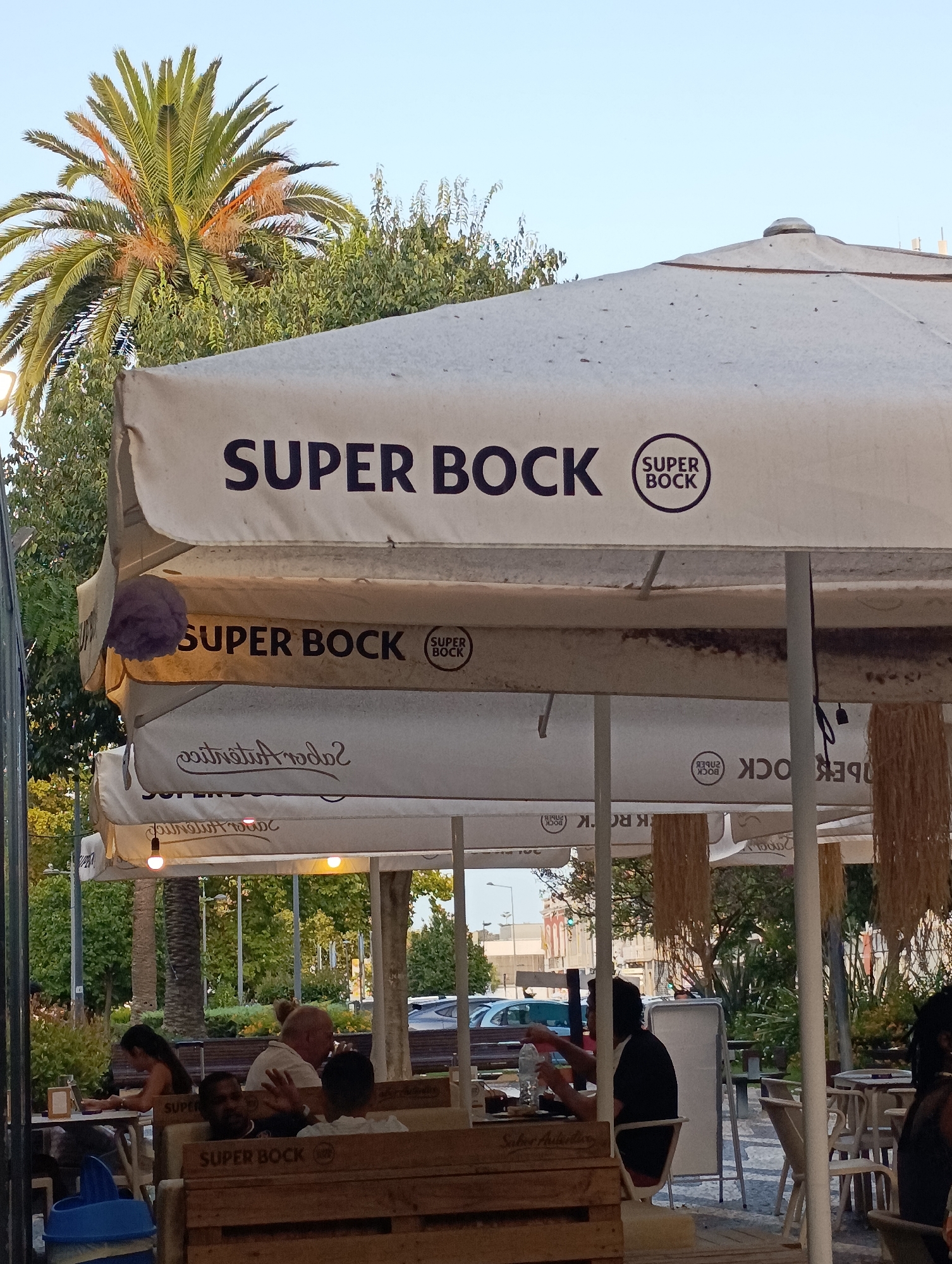 The image depicts an outdoor seating area of a cafe or restaurant, likely in Setúbal, Portugal, evidenced by the "Super Bock" branding, a popular Portuguese beer. Several layers of white canopies or umbrellas are visible, prominently displaying "SUPER BOCK" in large, dark lettering. Below the canopies, natural light illuminates the scene, casting shadows and highlighting the textures of the outdoor furniture. In the foreground, people are seated at tables, some interacting or looking towards the camera. One person is gesturing with their hands. In the background, lush green trees and a palm tree suggest a warm climate. The presence of a blue trash can and outdoor lighting fixtures indicate a functional urban setting. The overall atmosphere appears relaxed and casual, characteristic of an establishment catering to patrons enjoying the outdoors.