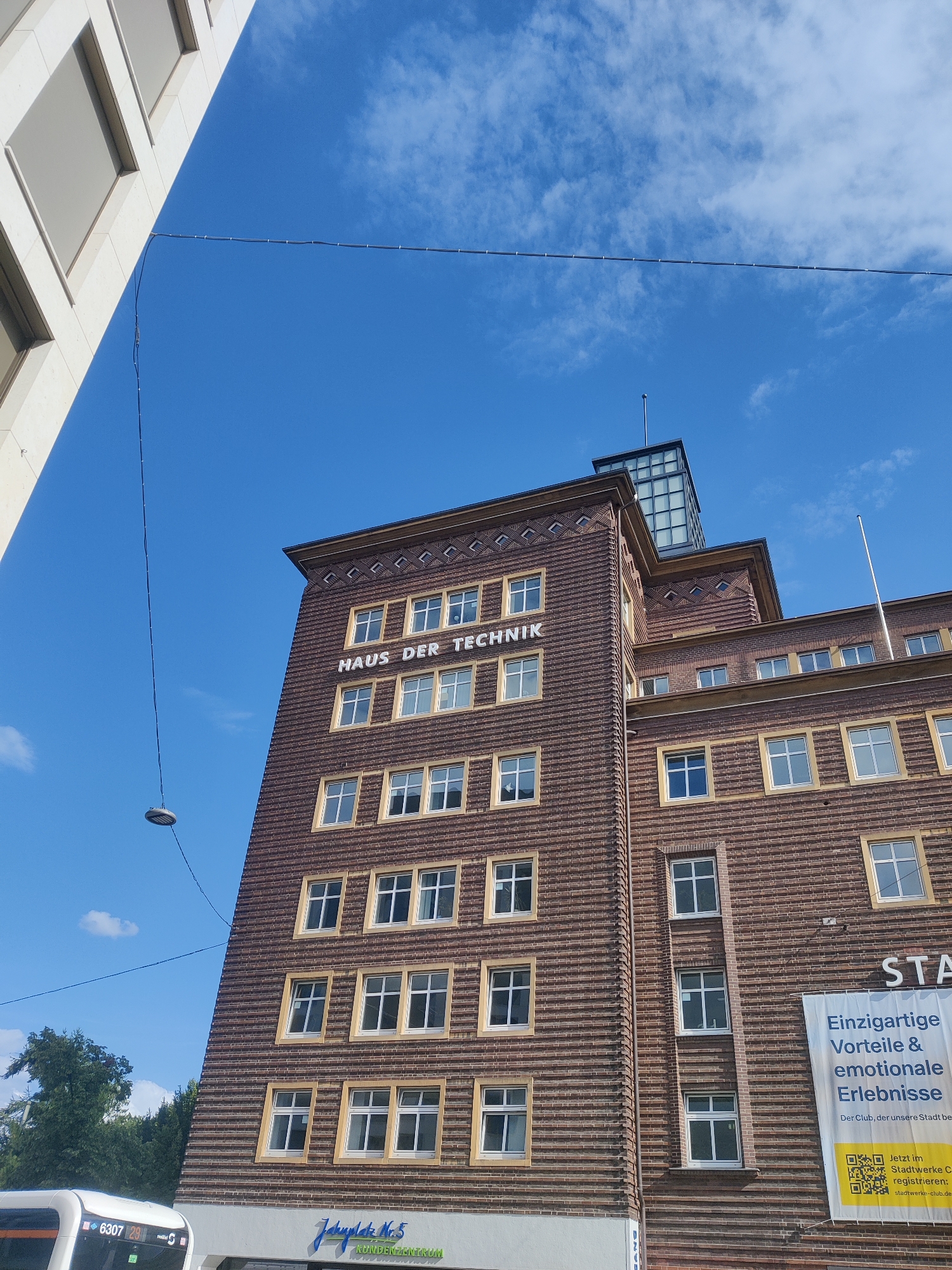 This is a photograph of the "Haus der Technik" building in Bielefeld, Germany, captured on a bright, sunny day with a clear blue sky dotted with a few wispy clouds. The building is a prominent, multi-story structure with a distinctive brick facade. The architecture features rows of windows with light-colored frames, arranged symmetrically on the brickwork. The name "HAUS DER TECHNIK" is emblazoned in large, white lettering across the upper section of the building.
At the very top of the building, there's a glass-enclosed structure, possibly an observatory or a modern addition, catching the sunlight. A flagstaff stands tall on the roof of a lower section of the building. A banner is displayed on the right side of the building, which contains text in German, including "Einzigartige Vorteile & emotionale Erlebnisse" (Unique advantages & emotional experiences) and instructions to register on "stadtwerke-club.de".
In the foreground, on the left, a portion of another building, clad in light-colored stone with rectangular windows, is visible, suggesting an urban setting. A street light hangs from a wire strung across the sky. To the lower left, the front of a bus is partially visible, displaying the number "6307 29" and the word "mobil". The overall scene suggests a bustling city environment on a pleasant day, likely during daylight hours.