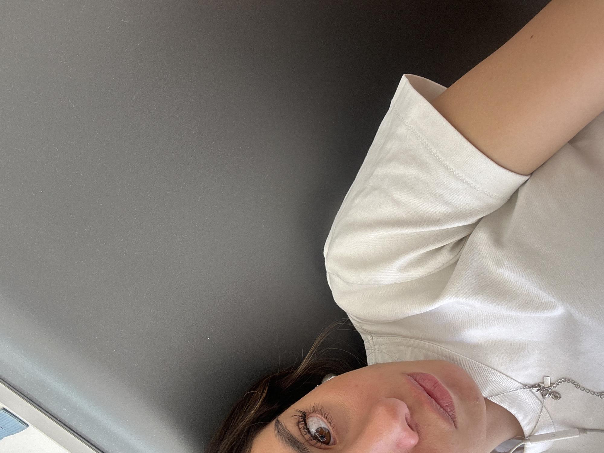 The image presents a close-up, tilted portrait of a person, likely female, captured from a slightly upward angle. The subject's face, including her right eye, nose, and mouth, along with a portion of her neck and shoulder, occupies the bottom right quadrant of the frame. She has dark brown hair, a brown eye with long eyelashes, and light pink lips. Her expression is neutral and calm.
She is wearing a light-colored, possibly off-white or cream, short-sleeved t-shirt. A delicate silver chain necklace with a small, circular charm is visible around her neck, resting on her chest. White earbuds or headphones are also present near her neck and ear.
The background, which makes up the majority of the image, is a solid, dark gray surface with a subtle texture and faint specks, possibly dust. This background shows a slight gradient, appearing marginally lighter towards the top left and darker towards the bottom right. The plain nature of the background offers no specific location cues, though it could be an interior wall, headboard, or a vehicle's interior. The lighting is soft and even, suggesting an indoor setting or an overcast environment. No visible text is present. The overall scene conveys a relaxed, casual moment, possibly a selfie taken while the person is resting.