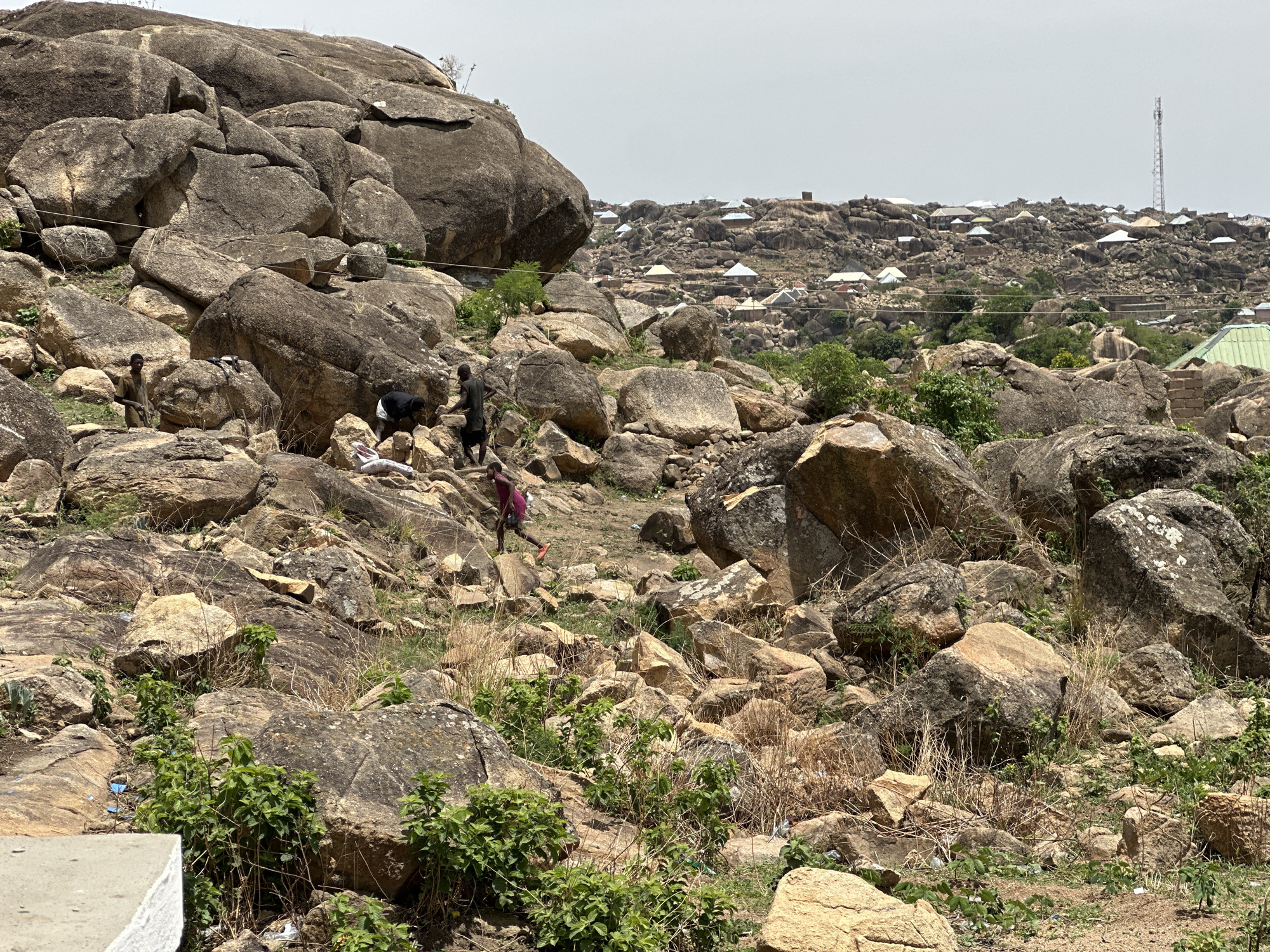 The image shows a rocky landscape near Jos, Nigeria, likely in or around a rocky outcrop region. Dominating the foreground and midground are large, weathered boulders and smaller rocks, interspersed with sparse vegetation. Several people are present among the rocks: one appears to be lying down, while others are standing or walking. The attire of the people suggests a casual, everyday setting. In the background, a cluster of buildings with diverse roof styles is visible, indicating a settlement. Further into the distance, a telecommunications tower rises above the houses. The sky is overcast, creating a muted, even lighting across the scene.