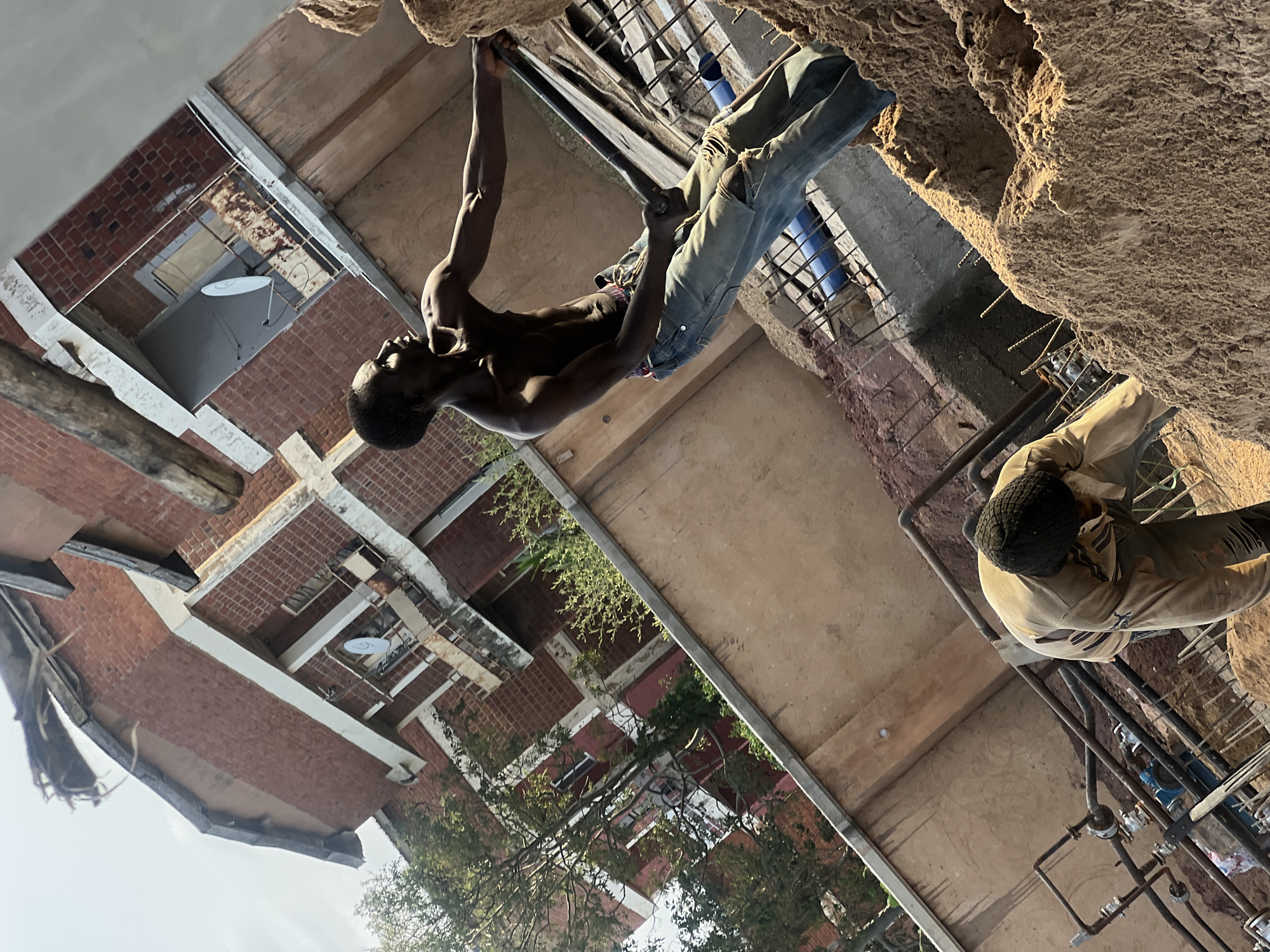 This image, set in Jos, Nigeria, captures a dynamic scene, likely at a construction site or an urban area undergoing renovation. In the foreground, a muscular, shirtless man with dark skin is suspended, performing what appears to be a pull-up or climbing motion. He grasps a horizontal metal bar (rebar) with both hands, wearing ripped blue jeans, and his head is tilted upwards in concentration.
To his right and slightly below, a second man stands on a lower level or scaffolding, viewed from behind. He wears a light-colored long-sleeved shirt and a dark knitted cap, appearing to observe or be engaged in work. The immediate surroundings consist of rough, unfinished surfaces of concrete or packed earth, with numerous exposed metal rods, pipes, and scaffolding suggesting active construction.
In the background, a series of multi-story brick buildings with white trim are visible, some appearing residential or commercial. A small white satellite dish is mounted on one of the structures. Patches of green trees and foliage are interspersed among the buildings, breaking up the urban landscape. The scene is brightly lit by daylight under a clear sky, casting noticeable shadows. No discernible text is visible in the image. The overall impression is one of physical exertion and ongoing work in a rugged, sunlit urban environment.