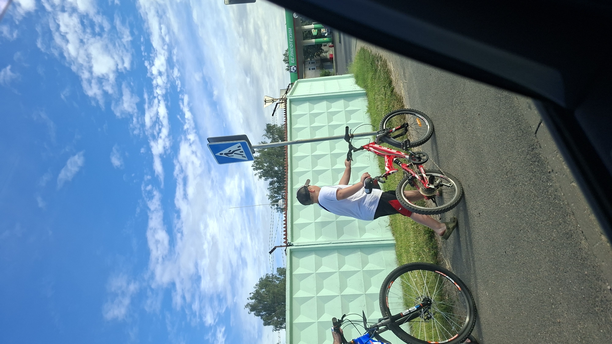 The image shows two bicycles and a young person in a white t-shirt and red shorts standing next to a pedestrian crossing sign. The person is holding onto the handlebars of a red bicycle. Another bicycle, mostly out of frame, is visible in the foreground. They are on a paved road next to a grassy area and a light green wall with a geometric pattern. In the background, there is a fence and trees, and a building with a green sign that reads "БЕЛНЕФТЕХИМ" (BELNEFTEKHIM) in white letters. The sky is blue with scattered white clouds, suggesting it is daytime with fair weather. The overall scene appears to be in an outdoor, possibly suburban or rural, setting. The pedestrian crossing sign indicates a designated area for people to cross the road. The presence of the bicycles suggests outdoor recreation or travel. The image is oriented vertically, with the sky at the top and the ground at the bottom. The overall mood is casual and summery.