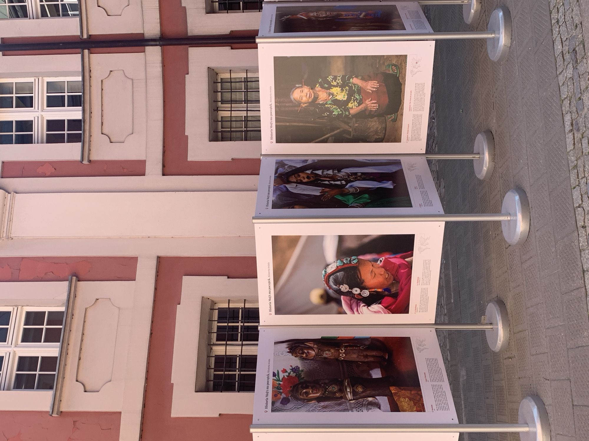 The image captures an outdoor exhibition of photographs in Poznan, Poland, likely on a sunny day given the strong shadows. The exhibition is set against the facade of a pink and white building with classical architectural elements, including multi-paned windows with white frames and decorative moldings. The building appears to be a historical structure.
In the foreground, several large, white display panels are arranged in a semi-circular fashion. Each panel features a photograph and text, suggesting a themed exhibition. The photographs showcased include portraits of people in traditional attire and possibly cultural artifacts.
Specifically, the visible panels display:
* The first panel from the left shows two wooden carved figures, possibly idols or masks, adorned with jewelry and colorful fabrics.
* The second panel features a close-up portrait of a young woman with elaborate headwear adorned with turquoise stones and silver coins. She is wearing a pink garment.
* The third panel displays a portrait of an elderly woman with distinctive ear adornments, wearing a colorful patterned shirt and a dark skirt. She is seated.
* The fourth panel, partially visible on the far right, shows another portrait of a person in traditional attire.
The display panels are mounted on silver poles supported by circular, silver bases resting on a paved ground composed of square and rectangular tiles. The overall scene suggests a public art installation or cultural event taking place in a pedestrian area of the city.
