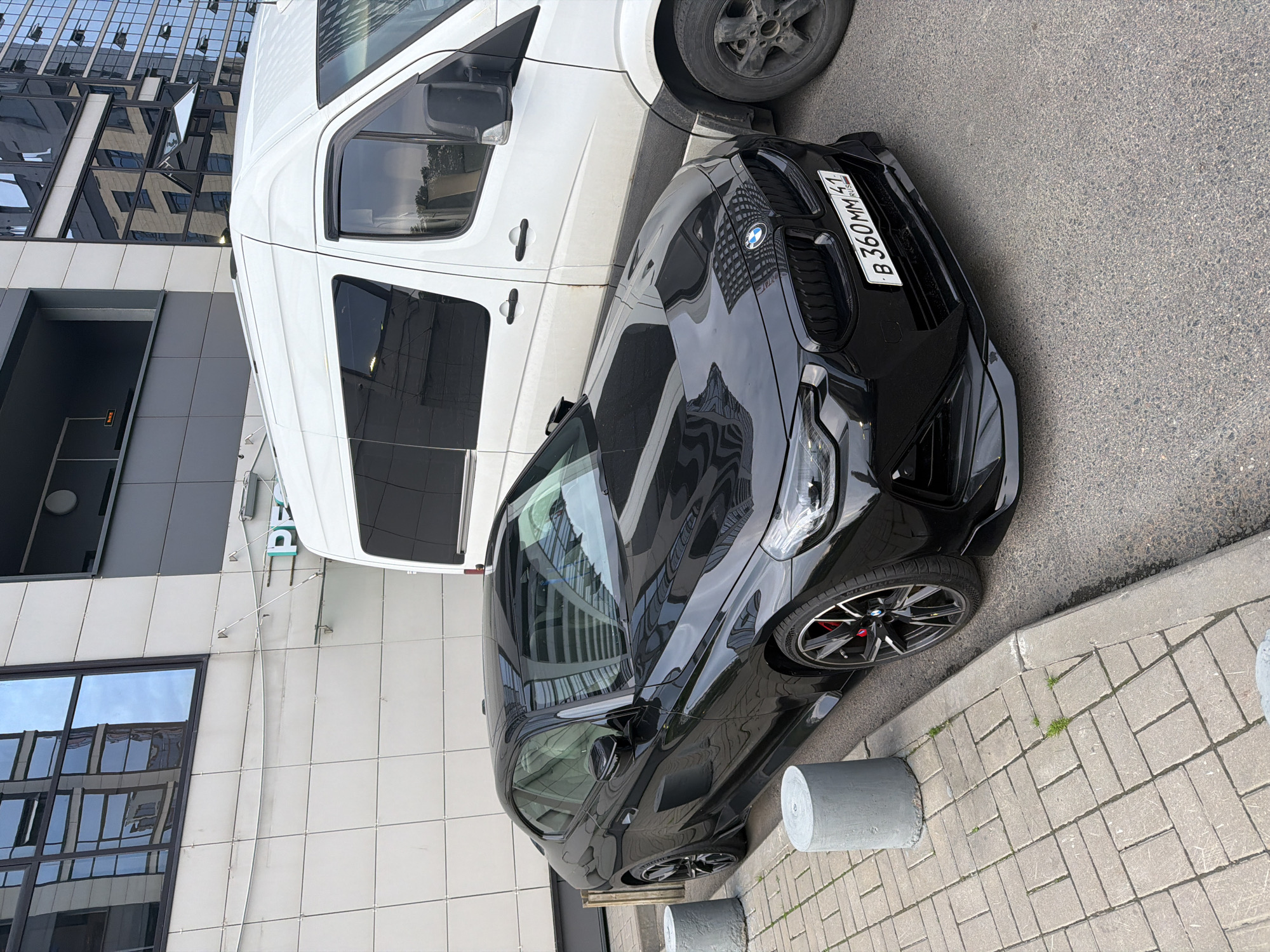 A black BMW coupe is parked next to a white van on a city street. The car is facing the camera and is slightly angled to the left. The van is parked to the right of the car and is partially obscuring the car's front passenger side. The car has a sporty design with a low profile and a large rear spoiler. The van appears to be a commercial vehicle, possibly a delivery van. The background shows a modern building with large windows. The street is paved with asphalt and there is a sidewalk on the left side of the image. The scene appears to be in an urban environment.
