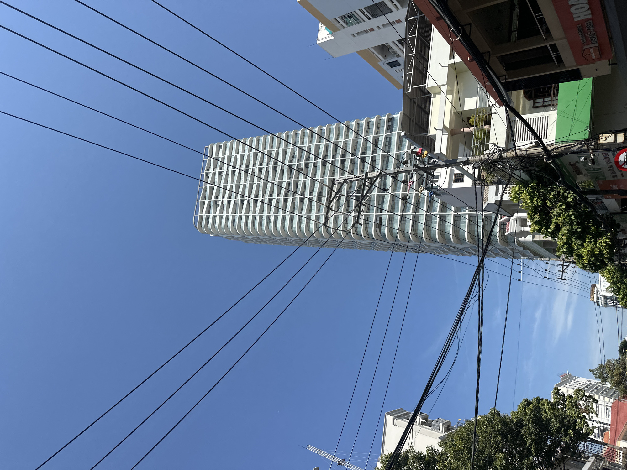 A low-angle shot captures a modern building with curved, white balconies set against a clear blue sky. The building's design is grid-like, with alternating sections of glazing. Multiple power lines stretch across the frame, converging near the building. Adjacent structures are visible, including a building with a green facade and an overhanging roof. Trees frame the bottom of the shot. A street sign and the tops of other buildings are visible in the background. The location is identified as being in Nha Trang Ward, Vietnam.