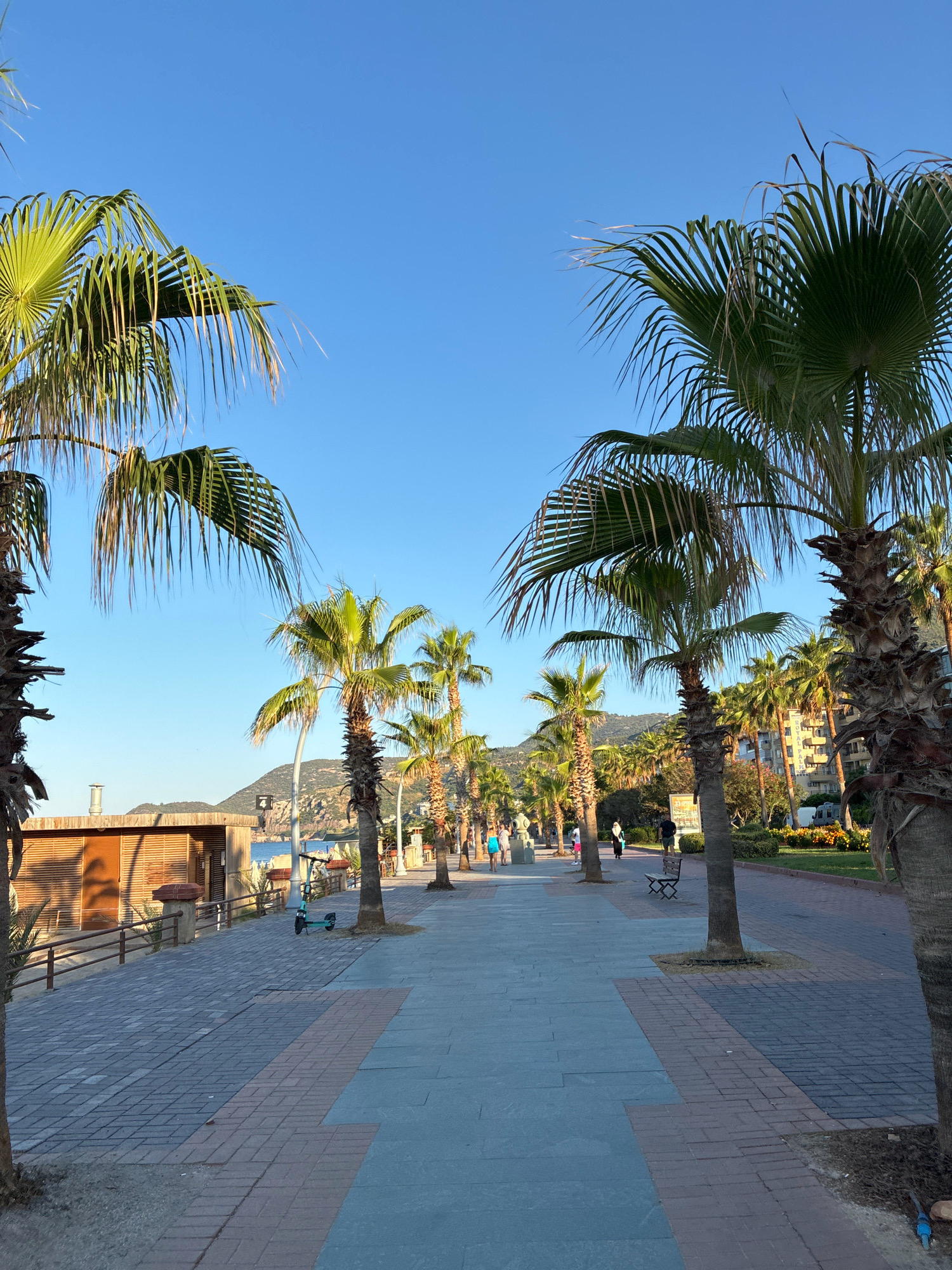 A paved promenade stretches into the distance, lined with numerous palm trees under a clear blue sky. The promenade features a walkway composed of rectangular light gray pavers with a decorative strip of reddish-brown bricks in the center, which is bordered by light gray pavers. To the left of the walkway, a sandy area is visible, separated by a low wooden fence and a wooden structure with louvered siding. Further down the promenade, several people are walking and a teal electric scooter is parked. In the background, the sea is visible, with a mountainous coastline and buildings beyond.