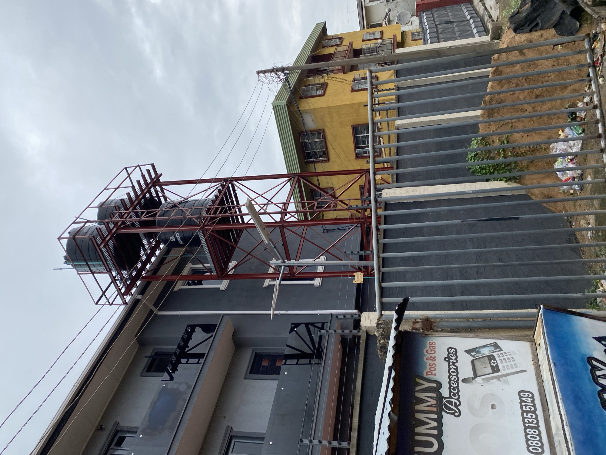 The image depicts an exterior scene in Abeokuta, Nigeria. A multi-story grey building is on the left, featuring balconies with glass railings and metal supports. To the right of the grey building, a red metal structure supports two large black water tanks on a platform. In the background, a yellow multi-story building with a green roof is visible. A grey metal fence runs across the foreground, with a pile of sand and some trash behind it. A sign with white and blue lettering, reading "UMMY Pos & Gas Accesories" and a phone number, is partially visible on the left side of the fence. Power lines are strung across the scene, and the sky is overcast. No people are present in the image.