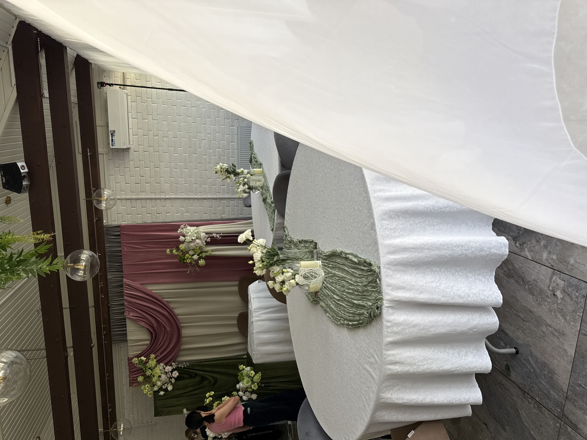 This image depicts an indoor event space, likely a banquet hall or restaurant, decorated for a celebration. In the foreground, a round table is covered with a white tablecloth featuring an embossed pattern. A green, crinkled fabric runner is placed diagonally across the table, adorned with two small white candles and a delicate white decorative element. A clear glass vase holds a bouquet of white flowers.
In the middle ground, other round tables are visible, also draped with white tablecloths. One table has a similar green runner and white floral arrangement. Further back, draped fabric panels in muted tones of green, beige, and pink create a backdrop. Two smaller round tables with white cloths are set against this backdrop.
To the left, a woman with dark hair pulled back is seen from behind, wearing a pink sleeveless top and dark pants. She is looking towards the floral arrangements.
The environment includes a brick-patterned wall, a white air conditioning unit mounted high on the wall, and hanging spherical light fixtures. The floor appears to be tiled with a gray stone pattern. In the far right of the image, a large expanse of white, sheer fabric dominates the frame, possibly a decorative curtain or drape. The overall impression is one of event preparation or a partially set-up celebration. The location is Votkinsk, Russia, suggesting a venue for local gatherings or events.