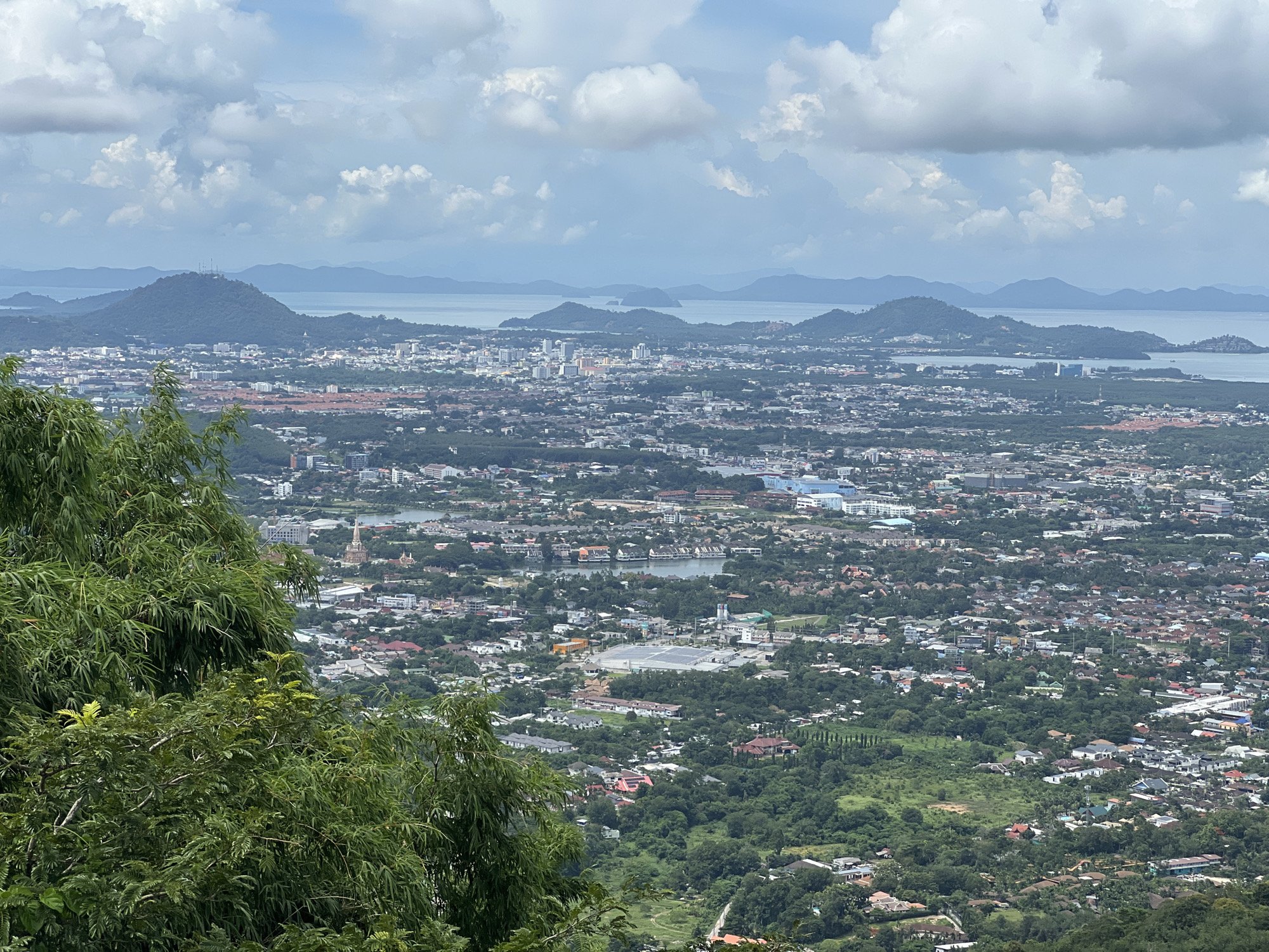 An aerial, wide-angle shot captures a sprawling city nestled between lush green hills and a distant, calm body of water under a cloudy sky. The foreground is dominated by the dense, vibrant green foliage of trees, partially obscuring the view.
Below, the cityscape is densely populated with a mix of buildings, ranging from low-rise residential areas with red-tiled roofs to mid-rise apartment complexes and occasional taller white buildings. Interspersed among the structures are pockets of dense greenery and several small bodies of water, possibly lakes or ponds. Further into the distance, the urban landscape gradually merges with the natural environment, leading to rolling hills covered in vegetation.
Beyond the city and hills, a wide expanse of calm, pale blue water stretches towards a hazy horizon, dotted with several small, dark green islands or peninsulas. The sky is a mix of bright blue and large, puffy white and grey clouds, suggesting an overcast but possibly pleasant day. The scene appears to be an overview of Karon, Thailand, looking out towards the coast. No people or specific actions are discernible in the image.