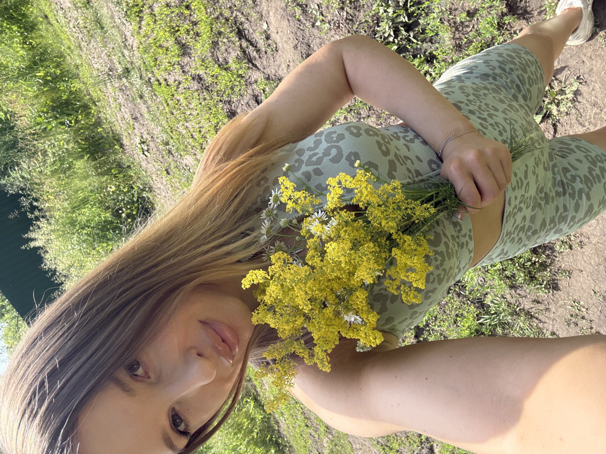 In this close-up, vertically oriented selfie, a young woman with long, blonde hair is captured outdoors. She is wearing a light green leopard-print sports bra and matching shorts. The woman is holding a bouquet of small yellow wildflowers and a few white daisies in her left hand, which rests on her stomach. Her right arm is bent, and her hand is not fully visible.
The background suggests a natural setting with dirt paths and green grass, hinting at a rural or park-like environment. Sunlight illuminates the scene, suggesting it is daytime, likely a warm and sunny day. The woman's expression is soft and relaxed, with a slight smile and her gaze directed towards the camera. A delicate silver bracelet is visible on her left wrist. The overall mood is peaceful and serene. The image was taken in Октябрьское, Russia. There is no visible text in the image.