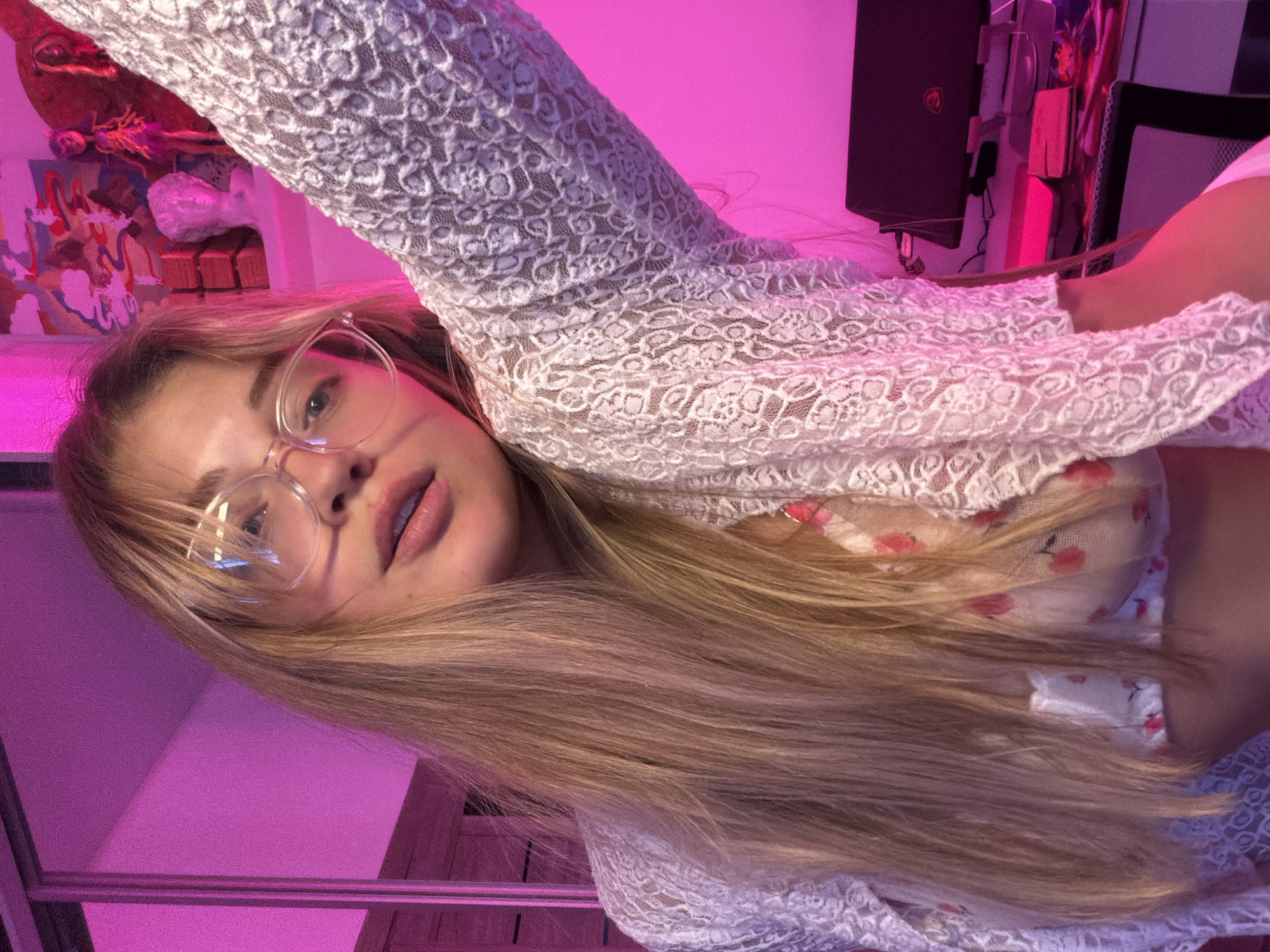 The image depicts a woman with long blonde hair and glasses. She is wearing a white lace top over a floral shirt. The background is illuminated with pink light. The location is not discernible from the image. There is no indication of the city of Большая Шильна, Russia.