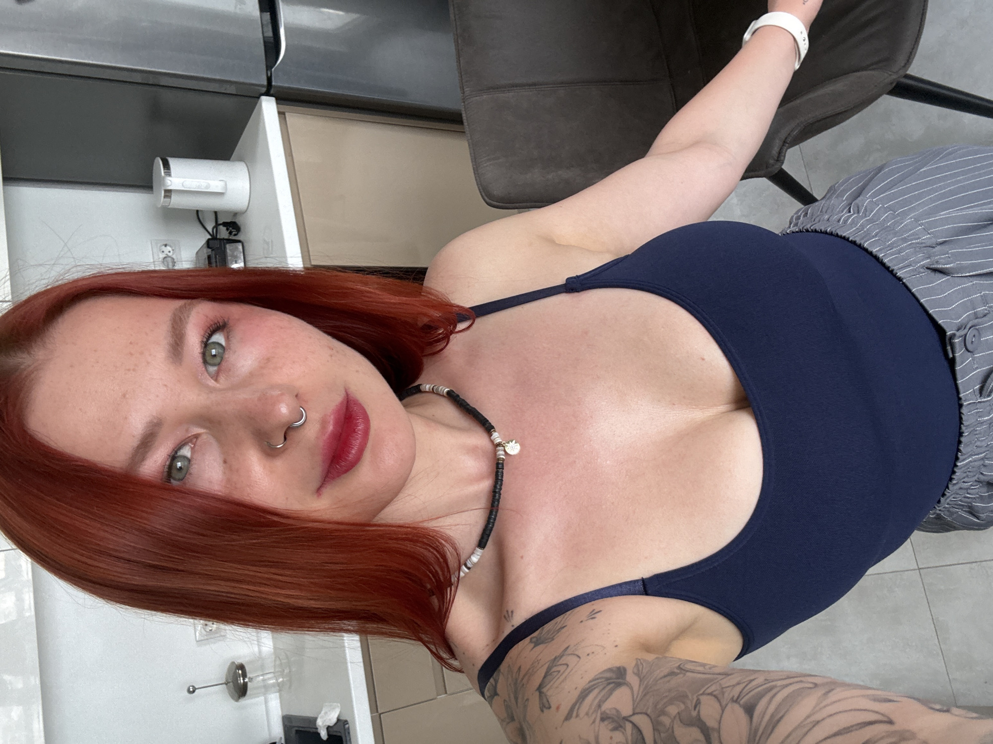 A woman with red hair and green eyes is angled to the right of the frame, wearing a dark blue tank top and striped shorts, sitting on a brown chair with a white watch on her left wrist. Her right arm is partially visible, showing a large tattoo. She has a beaded necklace and a double nose ring. The background includes kitchen appliances and cabinets. The location is most likely indoors, but I cannot confirm that it is in Rostov-on-Don, Russia, from the provided image.