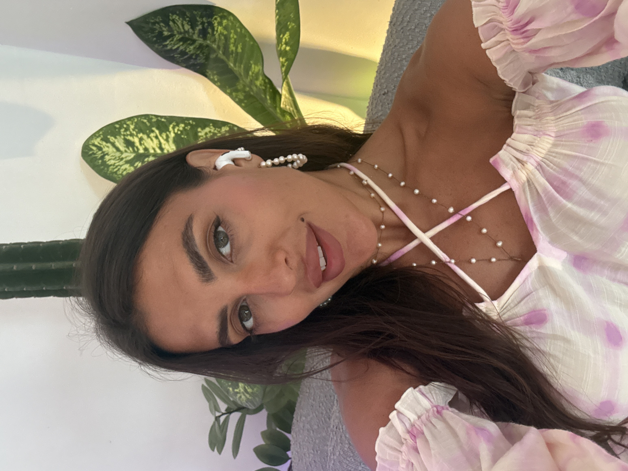 A young woman is depicted in a close-up, head-and-shoulders portrait, looking directly at the viewer with a subtle smile. She has long, dark hair parted to the side, framing her face. Her eyes are a light hazel or green. She is wearing a light-colored, possibly off-the-shoulder top with ruffled sleeves and abstract patterns in shades of purple and pink. Delicate, multi-strand pearl necklaces are draped around her neck and chest. In her visible left ear, she wears a white earbud and a decorative earring composed of small pearls.
The background features lush green foliage, including large-leafed plants with variegated patterns and a portion of a tall, columnar cactus. A light-colored wall is also visible behind her, with some natural light illuminating the scene. The setting appears to be either indoors or a shaded outdoor area, characteristic of a warm climate. The light suggests it is daytime. The woman's expression is pleasant and engaging. No text is visible in the image. The location context is North Kuta, Indonesia.