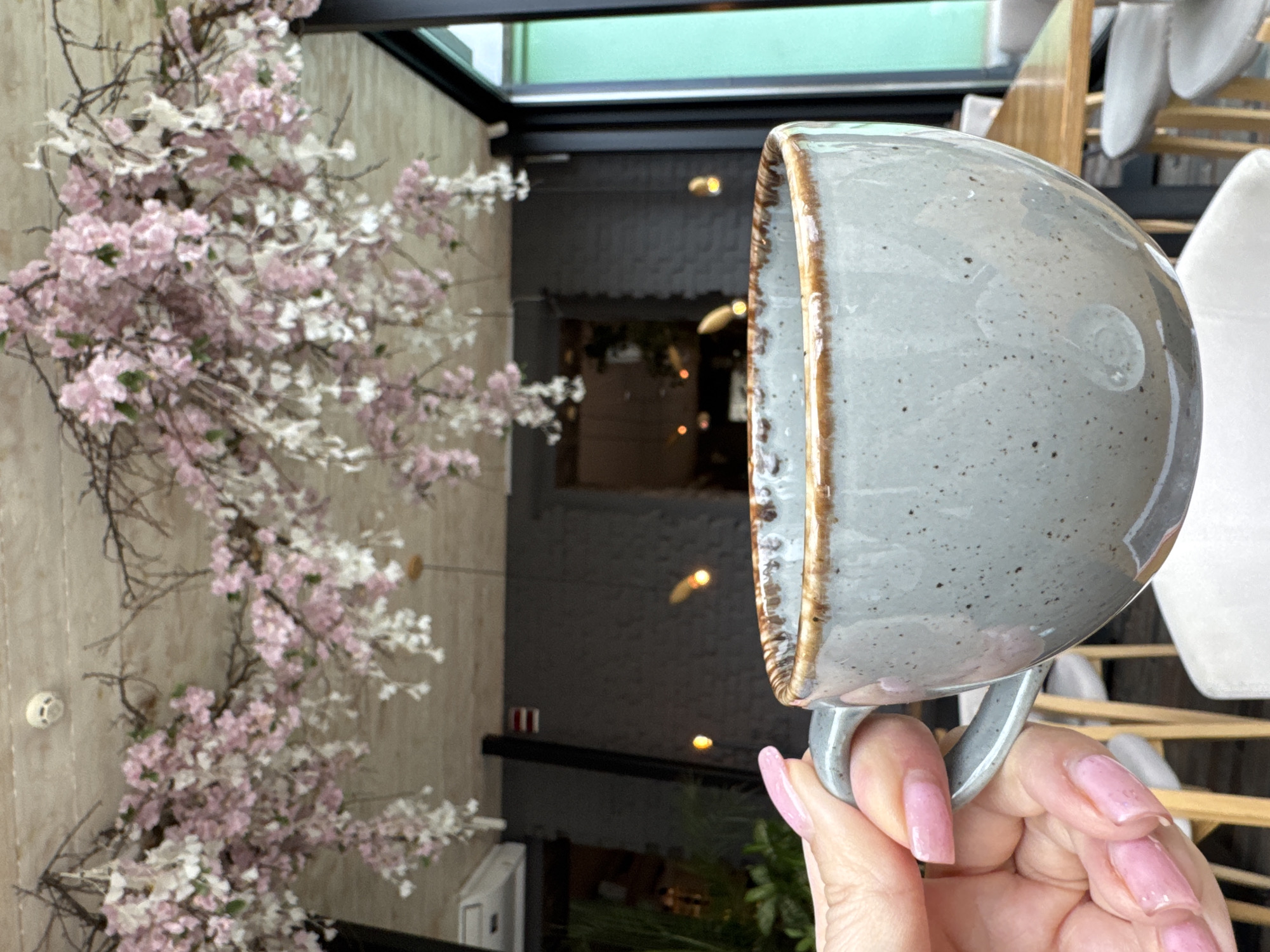 A close-up, medium shot shows a gray mug with brown rim held by a pink-nailed hand in front of a wall decorated with pink and white flowers and a dark-walled hallway in an indoor setting. The mug is held sideways with the opening facing left, revealing a textured gray surface with brown accents along the rim. The hand holding the mug has long, pink manicured nails. In the background, a wall covered in pink and white flowers is visible on the left, with branches extending towards the center. To the right of the flowers, a hallway with dark gray walls and a black ceiling can be seen. Small lights illuminate the hallway, hinting at a modern or minimalist interior design. Chairs and a table are visible in the background. The location is Unknown, Unknown.