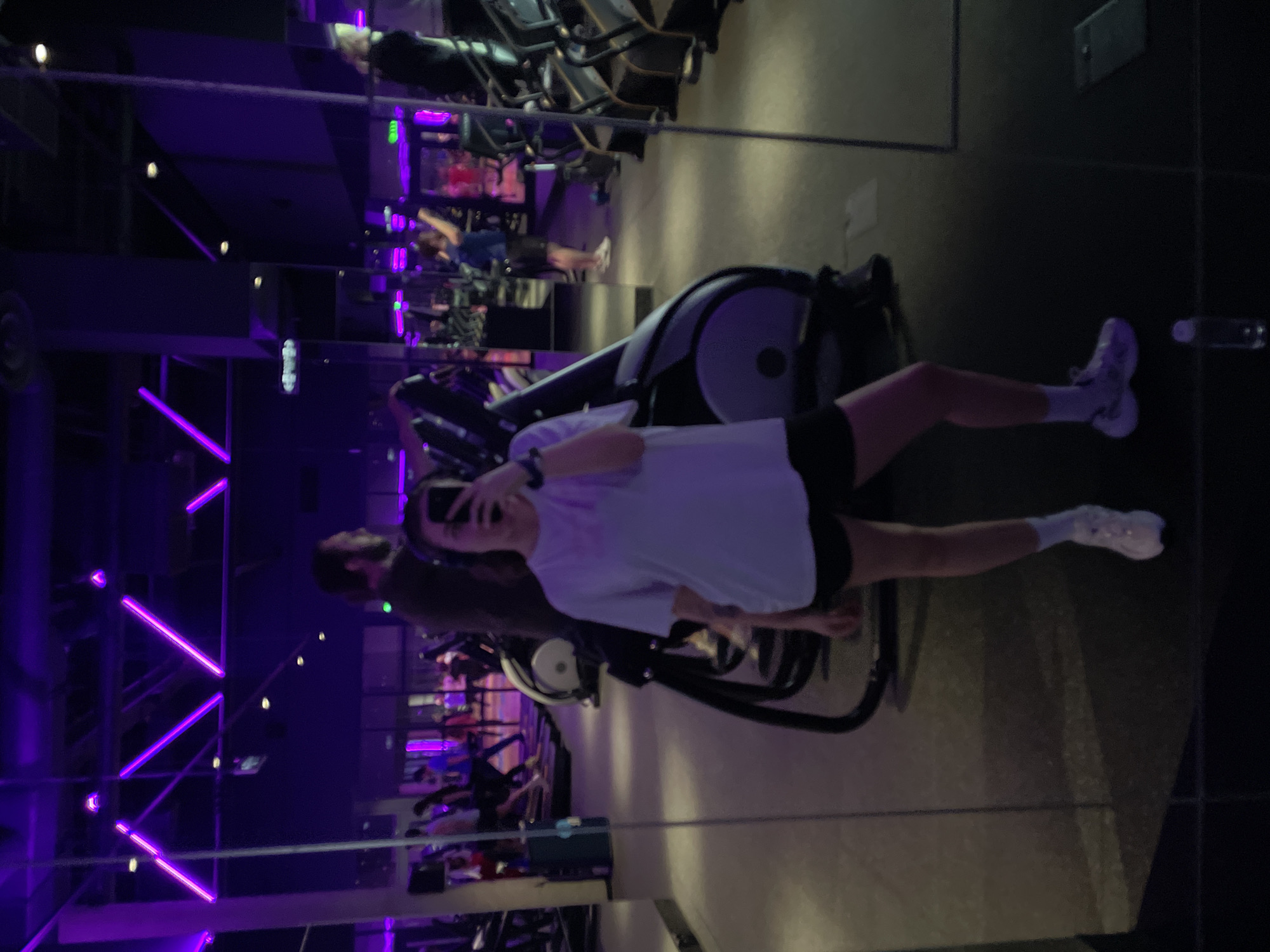 The image displays a mirror selfie taken by a person in a modern gym, identifiable by its distinctive purple lighting. The main subject, a young person, wears a light-colored short-sleeved t-shirt, black athletic shorts, white socks, and white athletic shoes. They are holding a black smartphone to capture their reflection, with a partial view of their face revealing a smile. The subject is positioned next to a piece of gym equipment, possibly a stationary bike or elliptical.
The gym interior is dimly lit, with the predominant illumination coming from neon-like purple LED strips arranged across walls and ceiling structures, creating a dynamic, modern atmosphere. In the background, reflected in the mirror, are numerous exercise machines, including rows of what appear to be treadmills and other bikes. The reflection also shows other gym patrons, with one individual, possibly male, with dark hair, visible in the mid-left background, also seemingly holding a phone. A clear water bottle with a white cap is visible on a surface to the right of the main subject's legs. The scene suggests an evening or nighttime workout session. Based on the provided context, the location is Moscow, Russia. No clear text is discernible within the image.