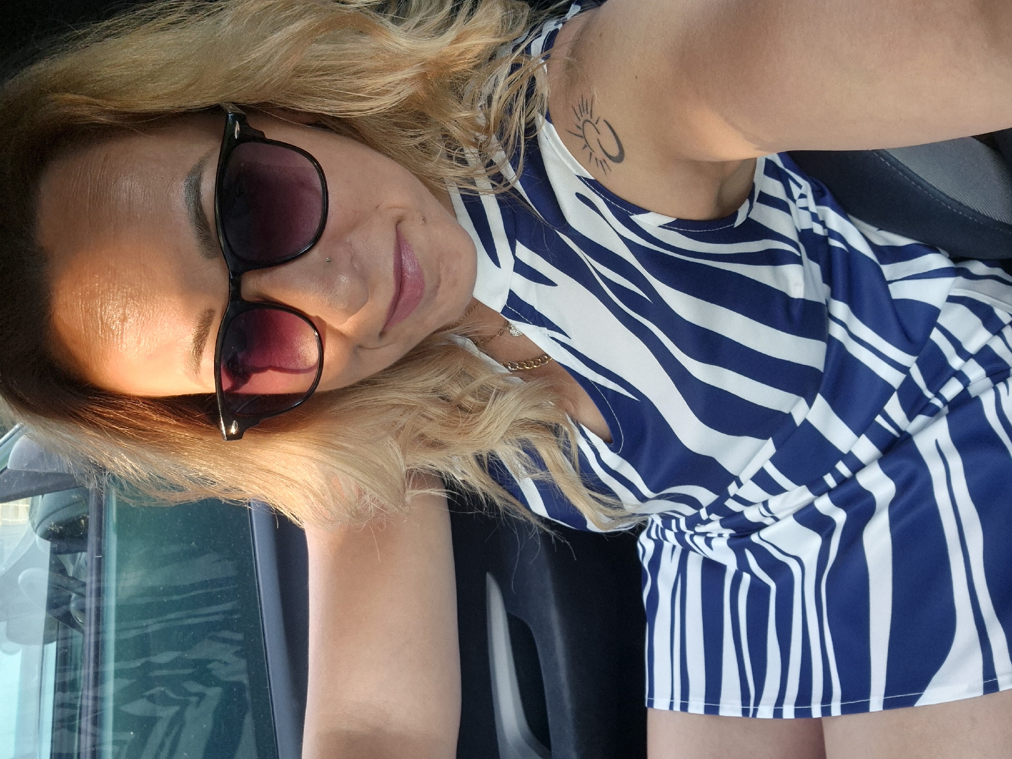 A woman is taking a selfie while sitting in the driver's seat of a car. She has blonde hair and is wearing sunglasses and a blue and white striped dress. She has a tattoo on her left shoulder. The car's interior is visible, including the dashboard and a portion of the passenger seat. The background shows the exterior of the car, with tinted windows and a glimpse of the surrounding environment, suggesting it might be daytime and outdoors. The image is taken from a slightly high angle, looking down at the woman.