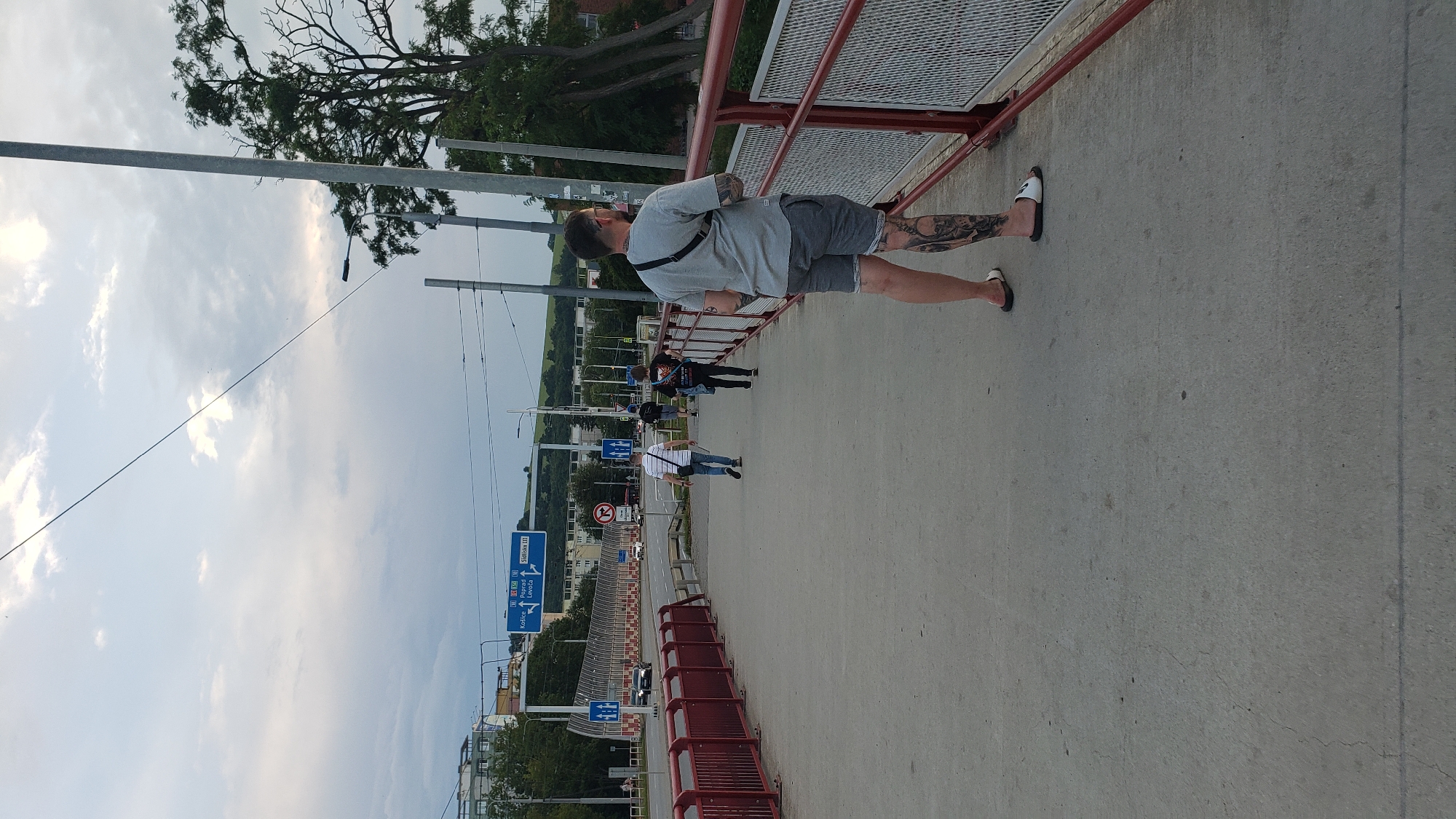 This image shows a pedestrian bridge in the city of Prešov, Slovakia. The bridge is made of concrete and has red railings on either side. In the foreground, a person with tattoos on their legs is walking away from the camera. In the background, there are several other people walking on the bridge, as well as cars on a road below. There are also trees and buildings in the distance, and the sky is cloudy. A blue road sign indicates directions to Košice, Levoča, and Prešov-Ši_o_kov_á.
