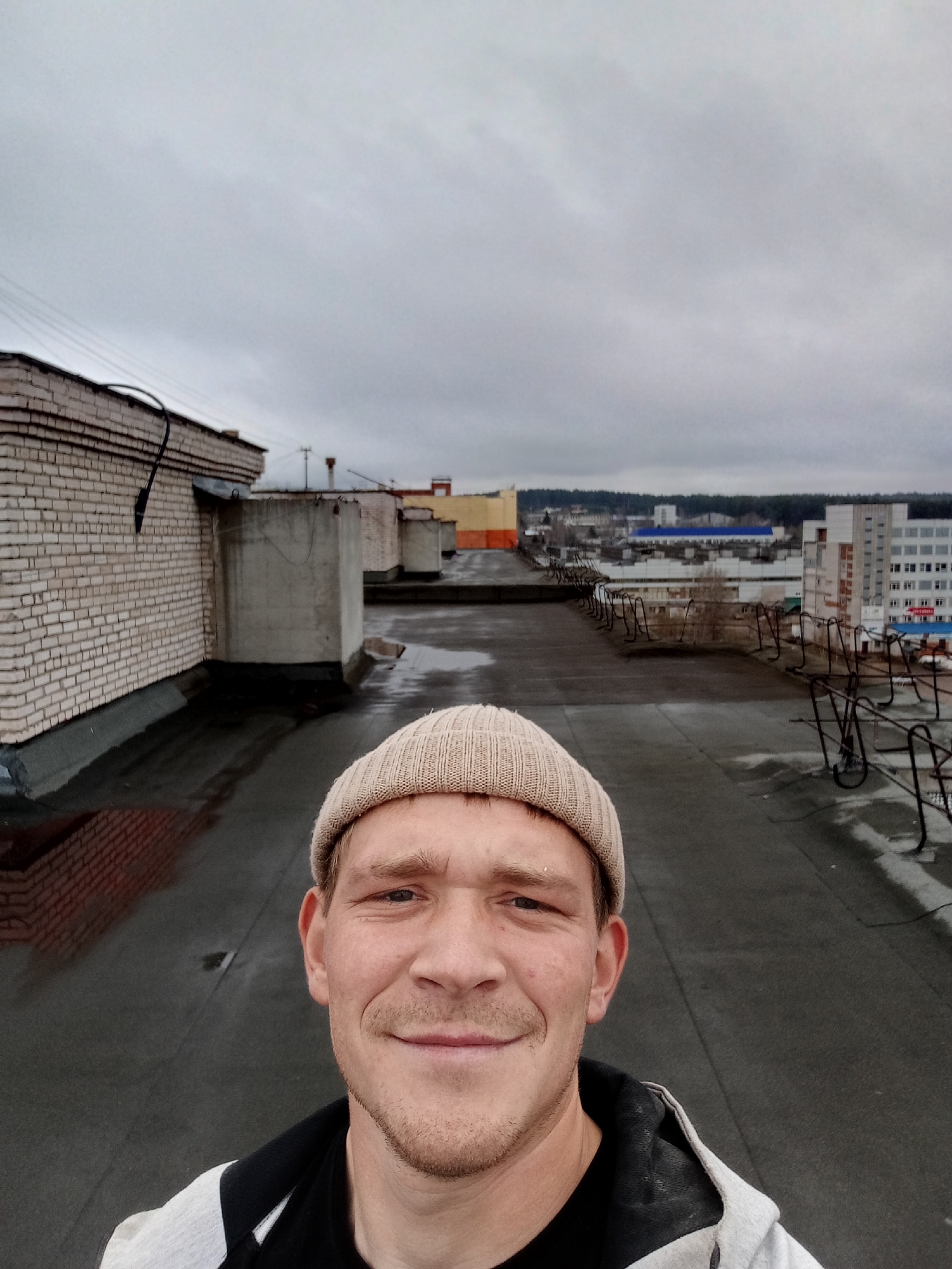 A fair-skinned man with a light beard and wearing a beige beanie is taking a selfie. He's on a flat rooftop, possibly of a building. Behind him, there's a brick building to the left and a cityscape extending into the distance under a cloudy sky. Based on the appearance of the buildings and environment, the location is likely Chaikovsky, Russia. There are metal security barriers/structures visible along the edge of the rooftop. The rooftop surface appears to be asphalt and has some visible water puddles.