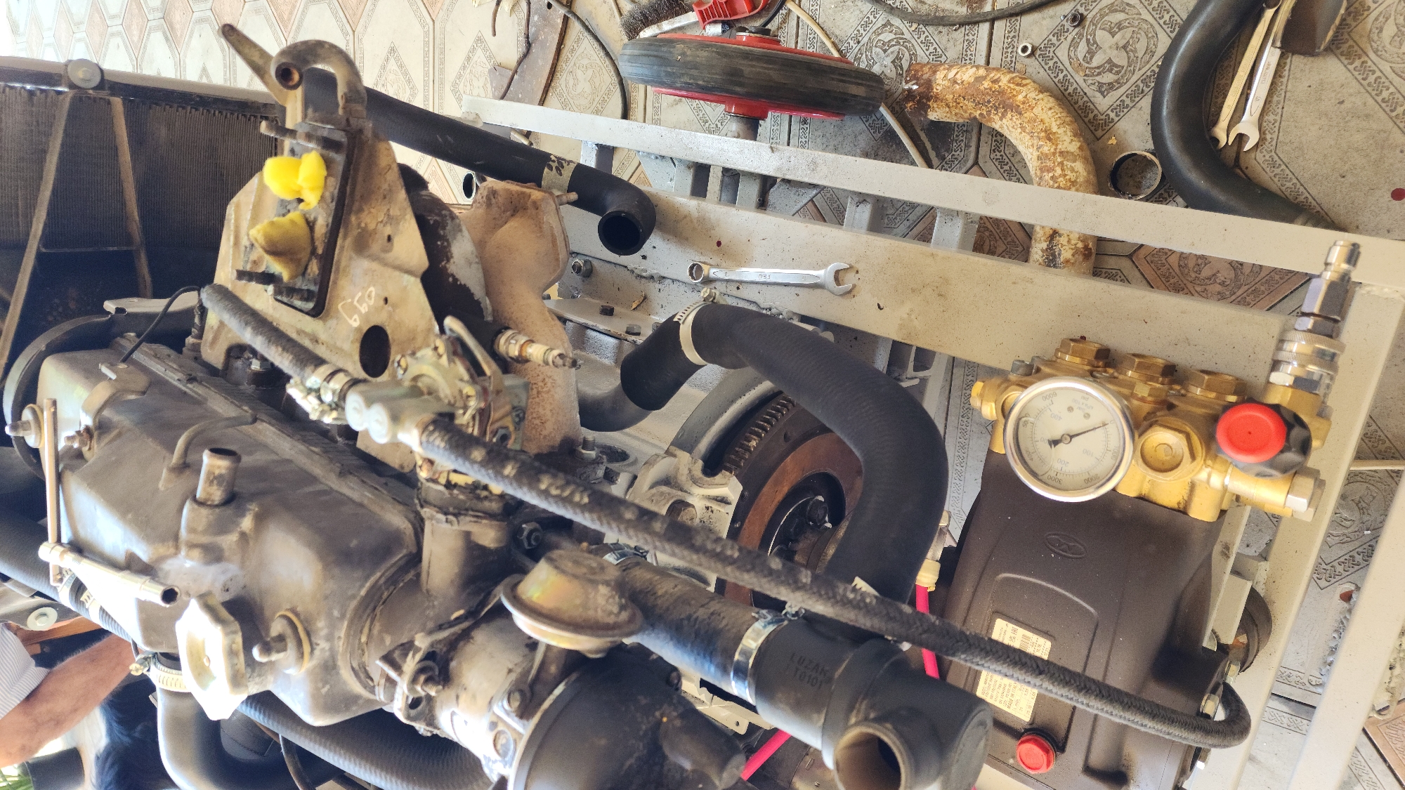 The image shows a close-up, low-angle view of a disassembled engine and pressure washer assembly on a tiled floor in Baghramyan, Armenia. The engine components, including the cylinder head, manifolds, and hoses, are visible and appear to be undergoing maintenance or repair. Several tools, including wrenches, are also present on the ground and near the engine. The pressure washer unit, with its gauge and various fittings, is positioned below the engine. The scene is outdoors, possibly a workshop or yard, with the tiled floor extending into the background. The lighting suggests it is daytime.