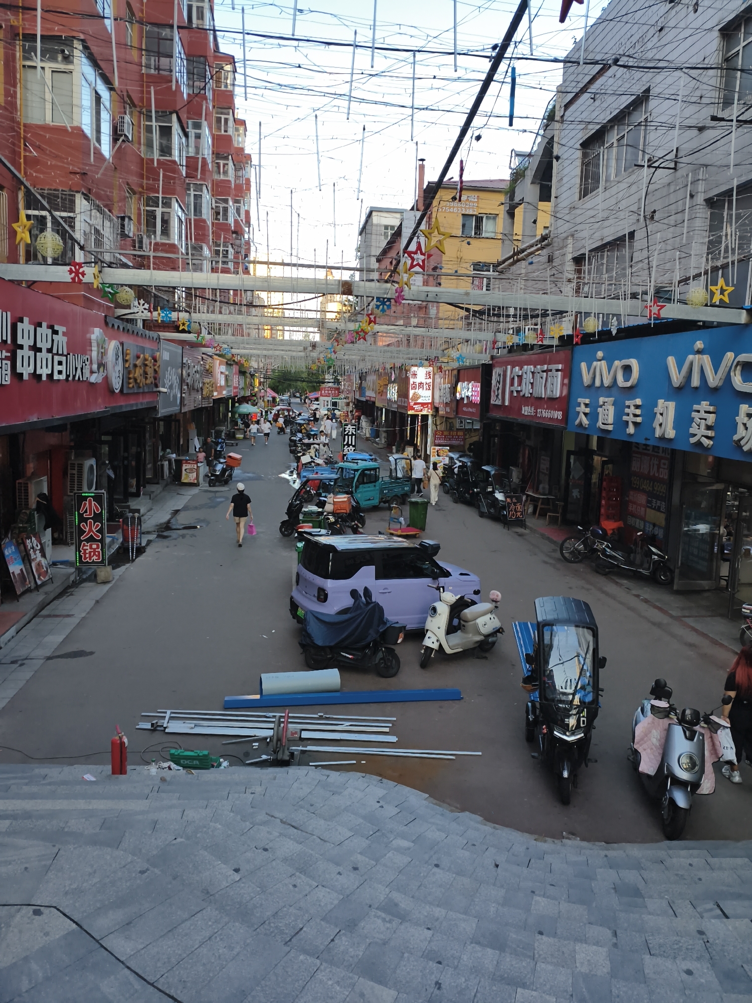 This image depicts a street scene in Mudanjiang, China. The street is lined with multi-story buildings adorned with various signs and decorations, including string lights and star-shaped ornaments. Several shops are visible, with signage in Chinese characters. A variety of vehicles, including electric scooters, three-wheeled utility vehicles, and a small purple car, are parked or in motion on the street. People are seen walking, and some are interacting with the vehicles. Construction materials, such as metal pipes and a green tool, are present on the ground near a set of stone stairs in the foreground. The sky above is clear, suggesting daytime.