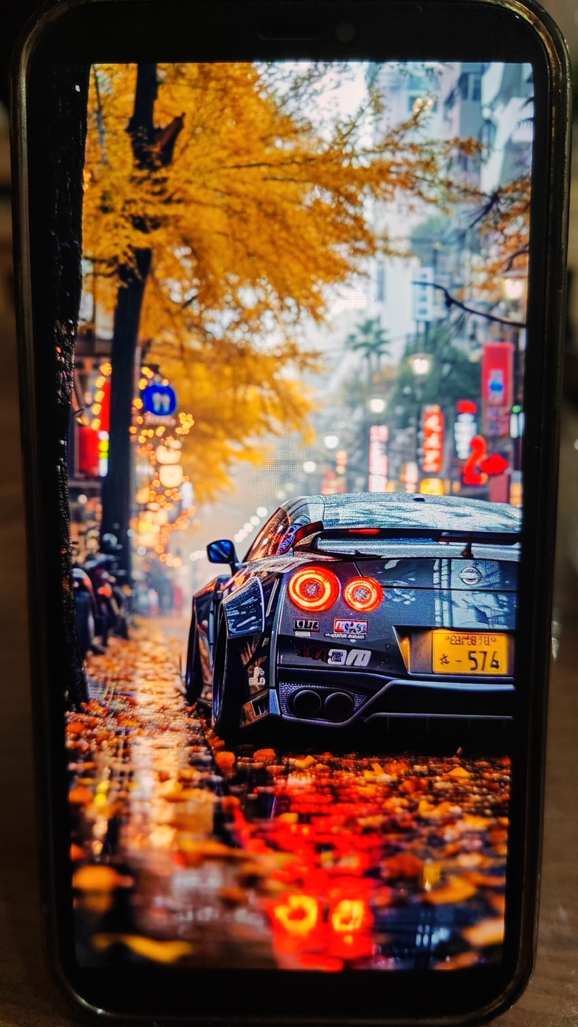 The image shows a phone displaying a wallpaper of a dark-colored sports car, possibly a Nissan GT-R, parked on a street lined with trees shedding golden-yellow leaves. The street appears wet, reflecting the colorful lights from buildings and streetlamps, suggesting it might have rained. The car's taillights are illuminated, casting a reddish glow. The background shows buildings with Japanese-style signage, indicating an urban setting. There are no people clearly visible in the image. The scene is evocative of autumn in a city, possibly in Japan due to the signage. There is no information in the image to suggest the location is Sochi, Russia.