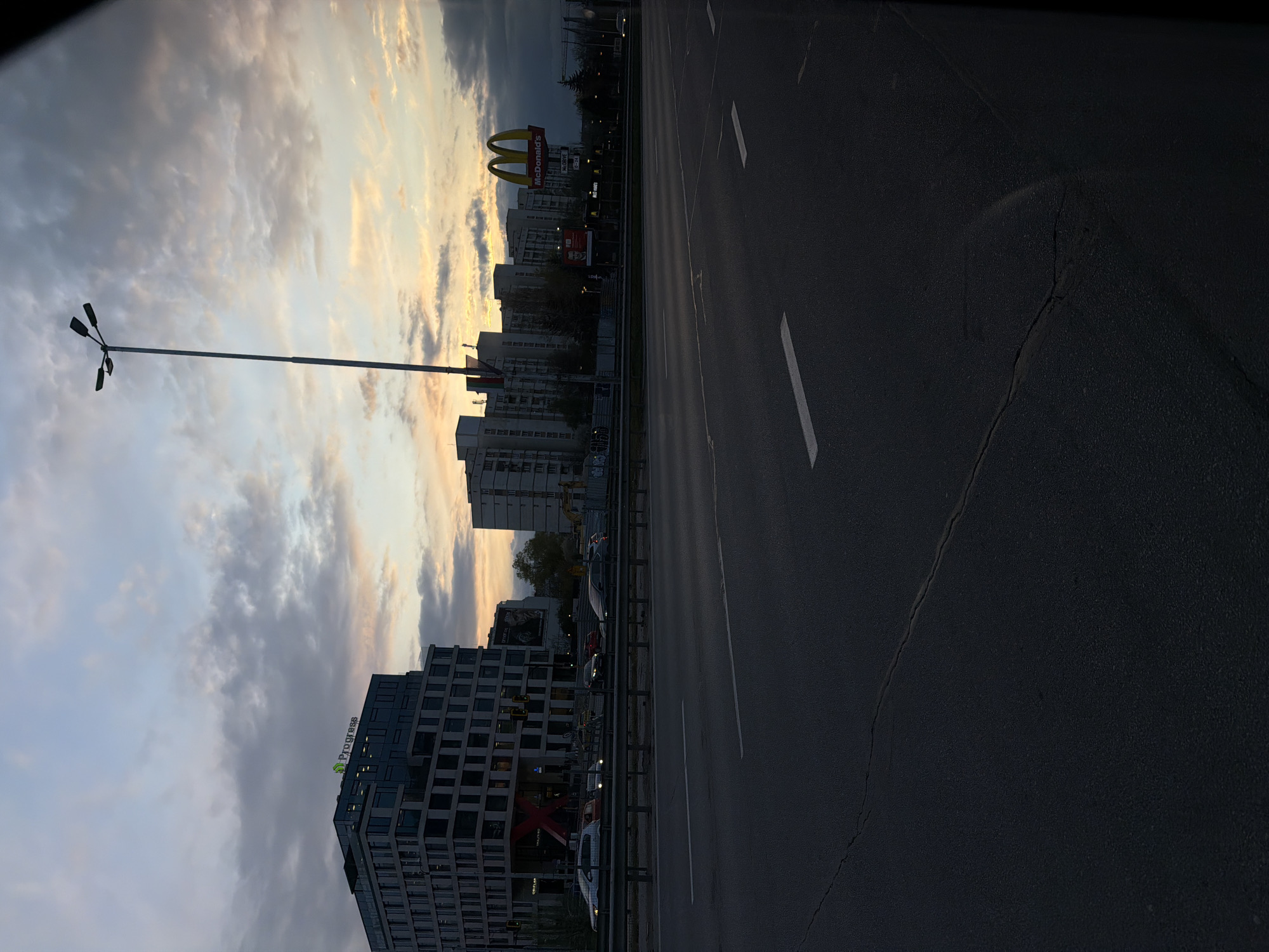 The image captures an urban street scene at what appears to be sunset or dusk. The dominant feature on the right is a multi-lane asphalt road with white lane markings, showing some cracks on its surface. A metal guardrail runs alongside the road, separating it from the buildings on the left.
The sky, occupying a large portion of the frame, displays a mix of conditions. On the far left, bright, warm orange and yellow hues suggest the sun setting below the horizon, while toward the center and right, the sky becomes overcast with darker gray and blue clouds.
Along the left side of the road, a series of commercial and residential buildings form a city skyline. A prominent McDonald's restaurant is visible with its large golden arch sign, advertising "DRIVE THRU" and "NON STOP 0-24", indicating it's open 24 hours. Further to the left, a modern building features "Progress" signage on its rooftop. Another building lower down has a distinct large red "X" and the text "ORIFLAME" visible. A billboard with the word "Epson" is also present.
A tall streetlight with three lamps extends into the sky from the left side of the image, positioned between the buildings and the road. Several cars are visible on or alongside the road, some parked, others possibly in motion, though none are clearly detailed. The overall atmosphere is that of a bustling urban environment transitioning from day to night.