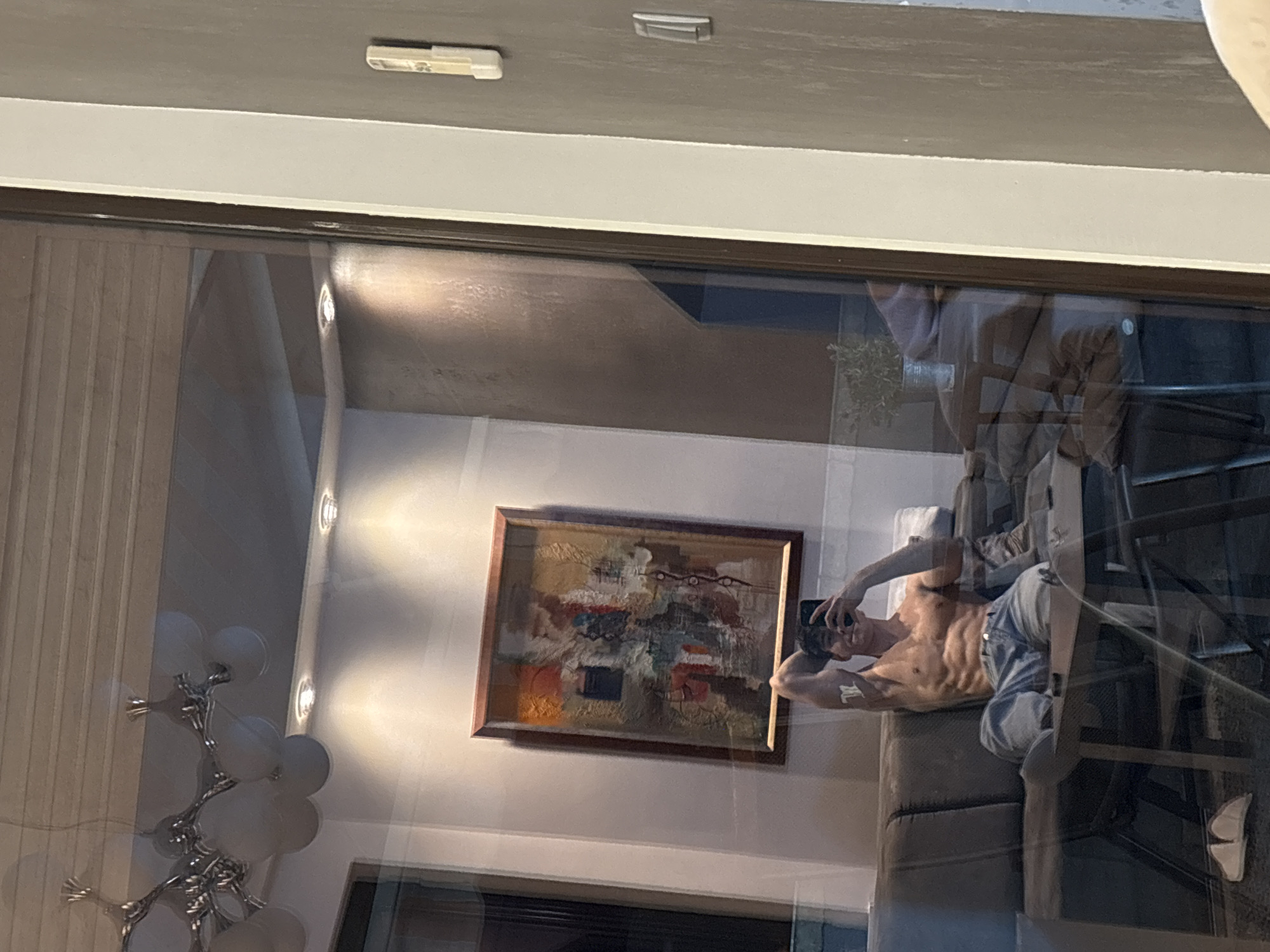 The image captures a reflection in a window or glass panel, revealing an interior scene, likely in Plovdiv, Bulgaria as per the provided context. The main subject is a muscular, shirtless man with well-defined abs, lying on a couch. He is wearing light-colored jeans or trousers and holding a smartphone, seemingly taking a selfie or video. A subtle mark or tattoo, resembling a letter like 'W' or 'M', is visible on his left bicep.
The reflected room appears to be a living area or lounge. On the wall behind the man, a framed abstract painting with a blend of reds, browns, blues, and other colors is visible. Above, several recessed spotlights cast distinct circles of light on the wall. To the left, a modern decorative floor lamp with multiple spherical white lights and a metallic, possibly chrome, base adds to the room's ambiance. Parts of other furniture, such as dark-legged chairs or a table, are faintly visible to the right of the couch. The ceiling in the reflection is light grey, with a white trim or frame at the top. The scene is illuminated by artificial indoor lighting, suggesting it might be evening or night. No explicit text is visible within the image, apart from the potential letter on the man's arm.