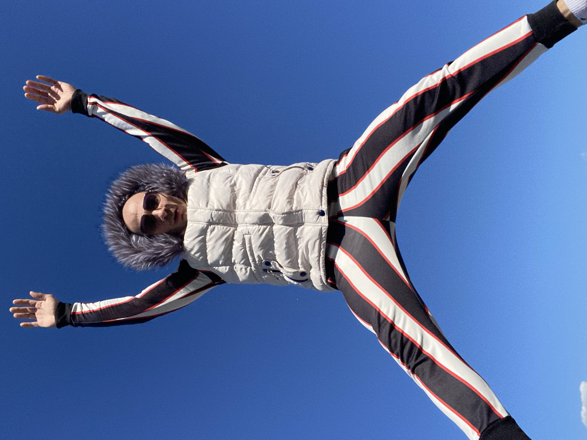 From a low-angle perspective against a clear blue sky, a person is seen with outstretched arms and legs. The person is wearing a light-colored quilted vest with a faux fur hood, and a striped black, white, and red tracksuit. The person is also wearing sunglasses. The location cannot be determined from this image.