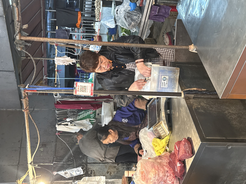 The image depicts an active market stall in Jiangjin, China. In the foreground, two women are engaged in sales activities at a dark metal counter. On the left, a woman with long dark hair, wearing a blue jacket over a patterned shirt, leans over a digital weighing scale, appearing to handle items in a clear plastic bag. Adjacent to her on the counter are multiple cuts of raw red meat and a large clear plastic bag containing smaller pieces of meat, alongside a yellow cloth. On the right, a woman with short brown hair, wearing a black leather jacket over a checkered apron, is actively reaching into a clear plastic container, seemingly packaging or processing items.
In the background, a man with a dark shirt or apron works behind a counter with a white basin. Overhead, a rudimentary structure of metal poles supports various hanging items including hooks, white and green plastic bags, colored towels, and a white QR code payment sign. An exposed light bulb provides illumination on the left side of the frame. Further in the background on the right, another market stall is visible with produce, possibly potatoes, displayed on a table. The environment is utilitarian, characteristic of an indoor or covered market.