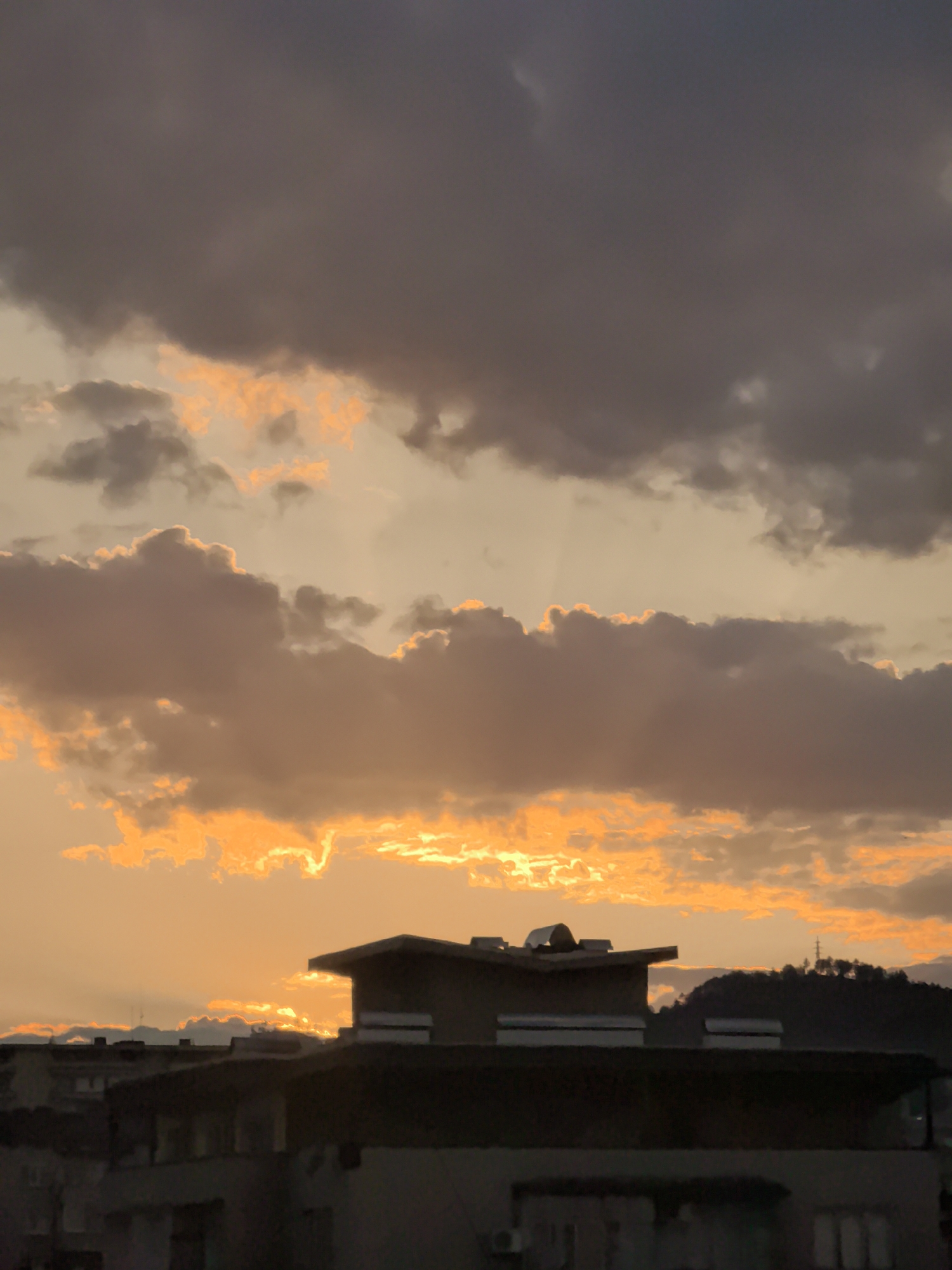 The image shows a sunset scene in what appears to be Stara Zagora, Bulgaria. The sky is filled with clouds ranging from dark grey to light orange, suggesting a sunset. Sunlight is breaking through the clouds, creating a vibrant, orange glow.
In the foreground, there are rooftops of buildings. The buildings appear to be residential. A hill is visible in the background, to the right, with a communications tower or pylon visible on its peak.