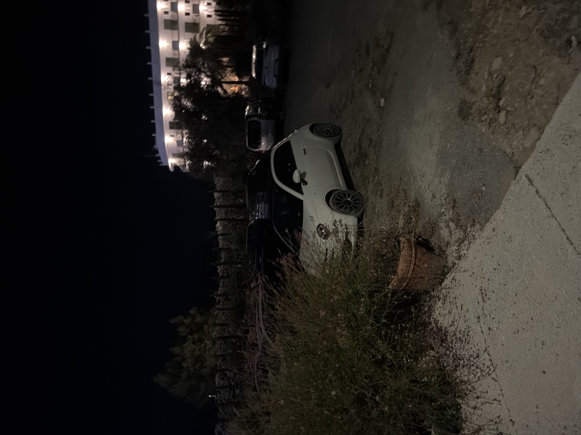 The image depicts a nighttime scene in Sant Antoni de Portmany, Spain, featuring a parking area and a building. A white Fiat 500 is parked in the foreground, angled slightly towards the right. Adjacent to the Fiat, a large bush with sparse foliage and some small pink flowers is visible, partially obscuring the car. To the left of the bush is a terracotta pot. In the background, several other vehicles are parked, including what appears to be a dark-colored car, a silver minivan, and a light-colored sedan. Behind the parking area, a multi-story building with illuminated windows and balconies is visible, likely a hotel or residential complex, with trees in front of it. The ground in the parking area is a mixture of gravel and dirt. The overall lighting suggests artificial illumination from the building and possibly streetlights.