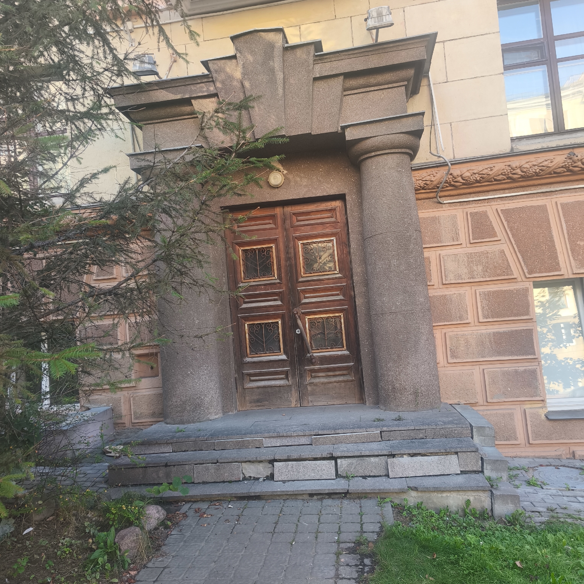 The image shows the entrance of a building with a double wooden door, featuring four small square windows with ornate metal grills on each door. The entrance is flanked by two large, rough-textured columns and is accessed by a set of stone steps. The building's facade is adorned with decorative stone elements and a textured wall. A large evergreen tree partially obscures the left side of the entrance. The surrounding area includes a paved walkway and some greenery. The location is identified as Minsk, Belarus.