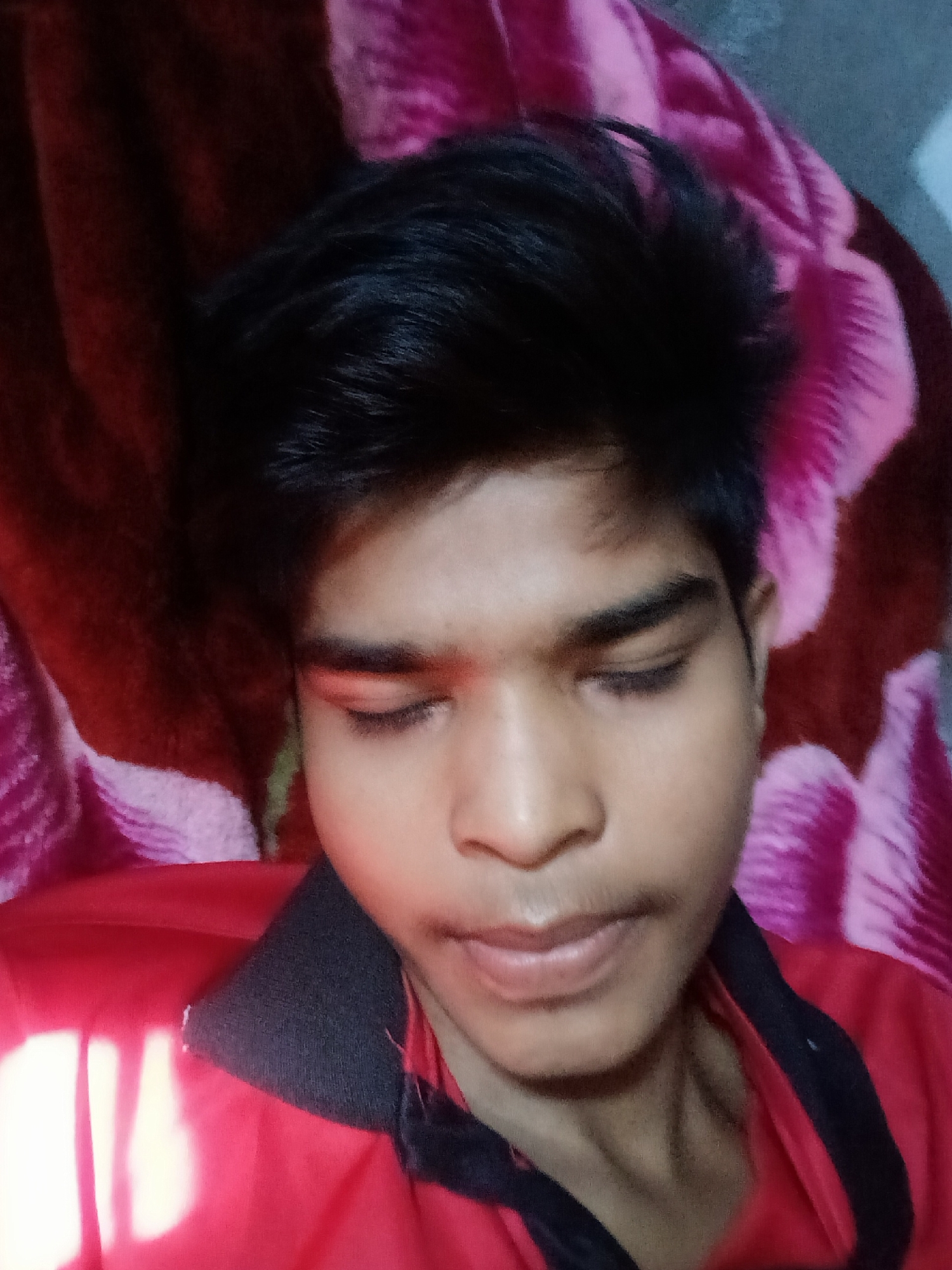 The image shows a close-up of a young South Asian male with dark hair and brown eyes, who appears to be lying down. He is wearing a red shirt with a black collar. The background consists of a pink and dark red patterned fabric, possibly a blanket or bedspread. The lighting suggests an indoor setting, possibly a bedroom. There is no identifiable location within the city of Bhopal, India.