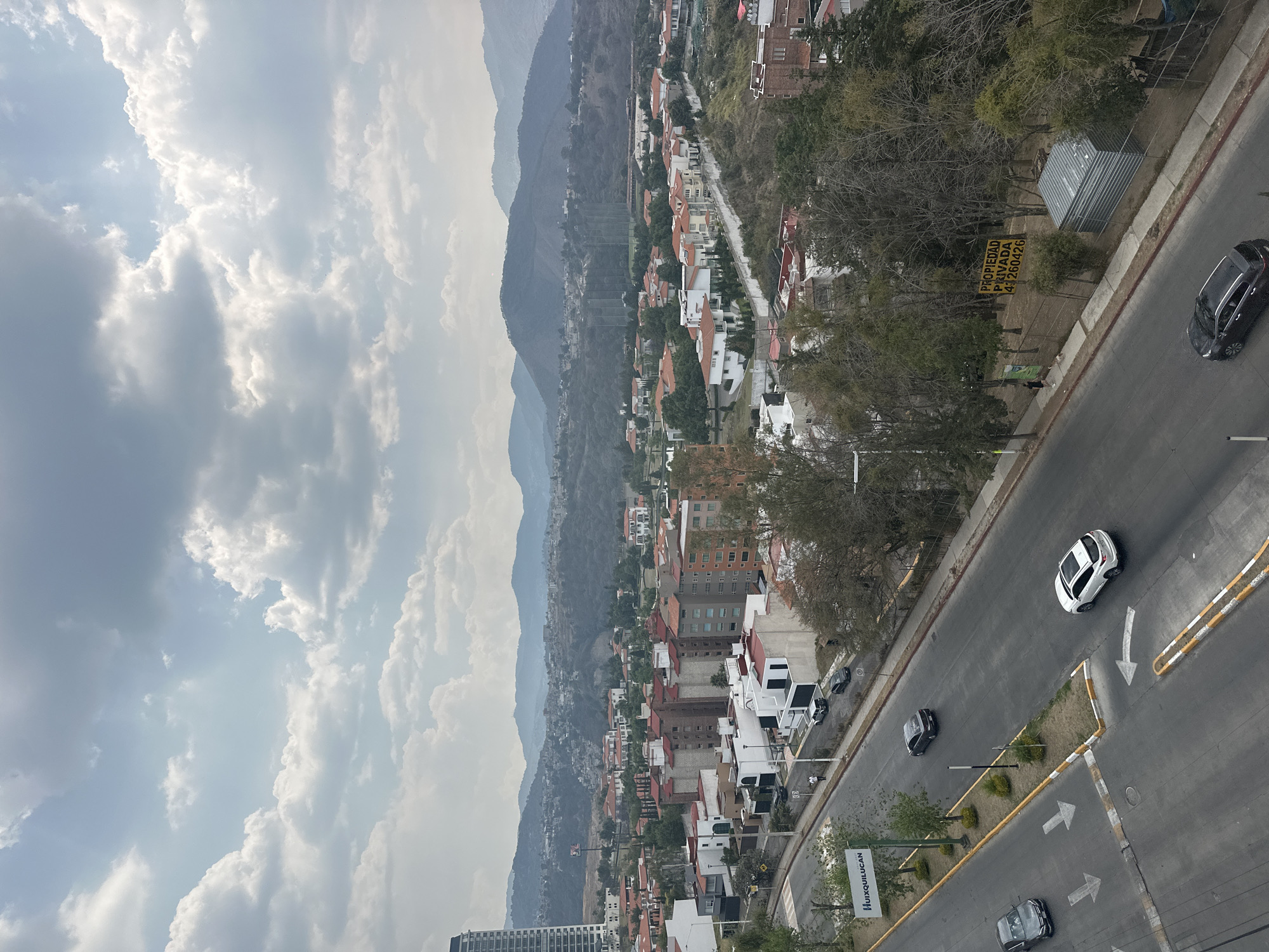 The image presents an elevated view of Jesús del Monte, Mexico, characterized by a mix of urban and mountainous scenery. The sky is filled with numerous clouds.
In the foreground, a road is visible, with several vehicles traveling on it.
The midground features densely packed buildings with red rooftops. Beyond the buildings, mountain ranges appear in the distance, adding depth to the landscape.