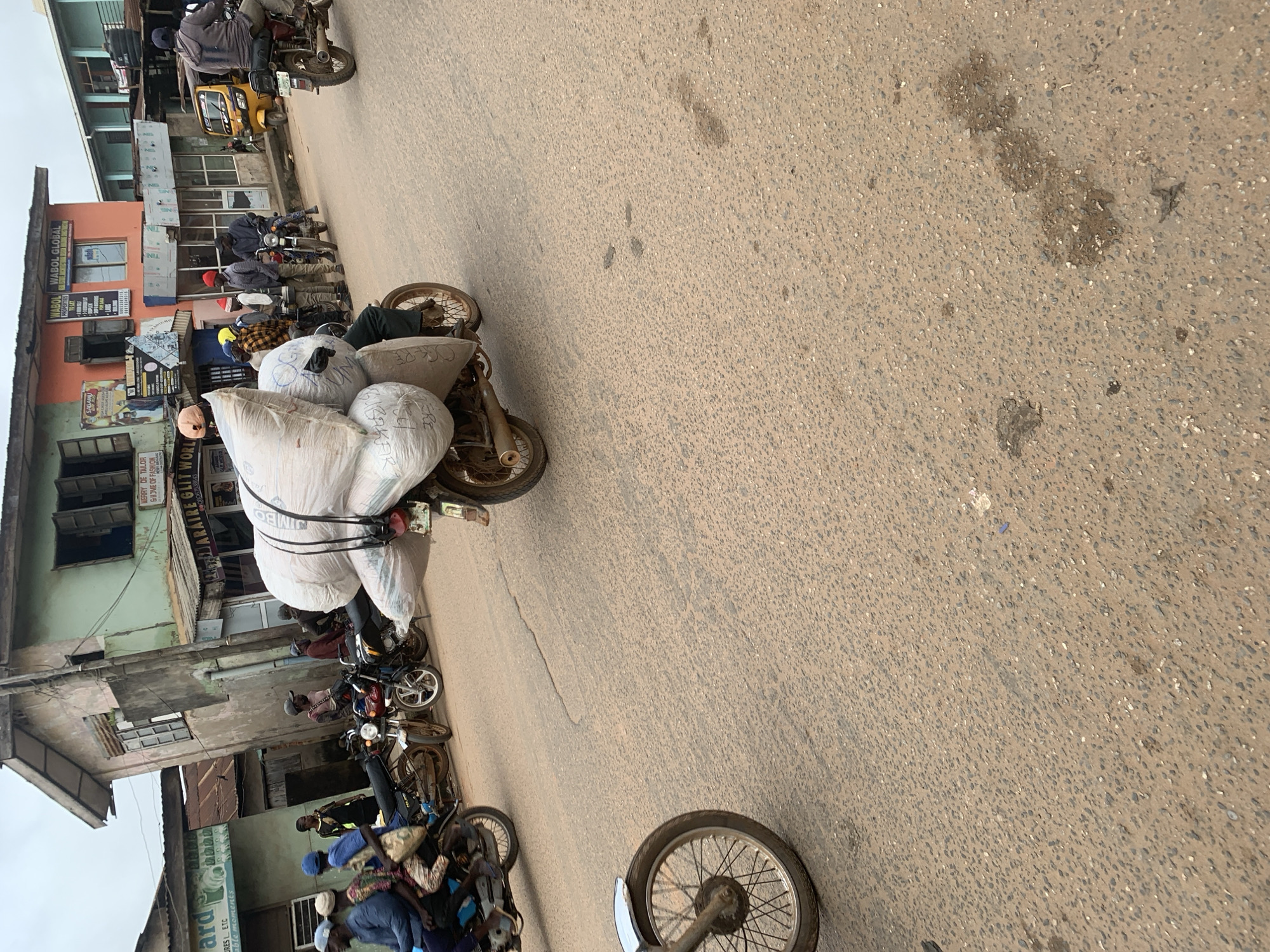 The image depicts a street scene in Shagamu, Nigeria, with a focus on motorcycles and their riders. In the foreground, a motorcycle is loaded with large sacks of goods, obscuring the rider's legs. Another motorcycle with a prominent front wheel is parked on the left, with its wheel and part of the frame visible. In the mid-ground, several motorcycles are parked or in motion. One motorcycle is carrying a rider who is leaning forward, also with large sacks secured behind them. Other motorcycles are visible with riders and passengers. The background features buildings, including a multi-story structure with an orange and green facade and other commercial buildings. A yellow three-wheeled vehicle is also present on the right side of the road. The road surface is paved and appears somewhat worn. The sky is overcast. The overall scene suggests a busy commercial area where motorcycles are a primary mode of transportation and delivery.