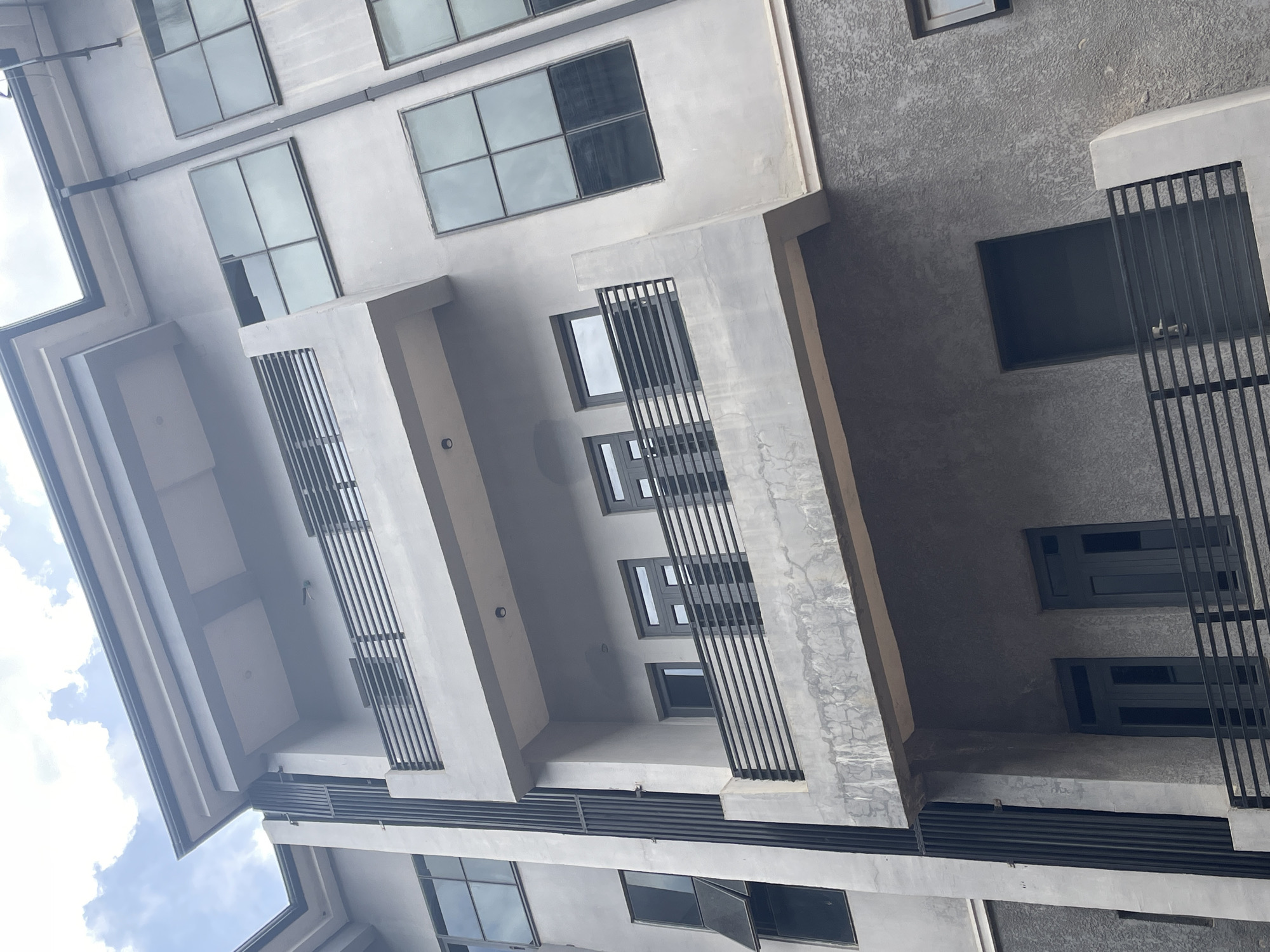 This is a low-angle, exterior, medium shot of a modern, multi-story building in Abuja, Nigeria. The building's facade is predominantly gray, with a textured finish on the lower levels and smooth concrete on the upper levels. Several balconies with black metal railings are visible, as are numerous windows with dark frames, many of which are reflective. Some of the windows are open. The building features multiple levels, each with balconies and windows, suggesting it might be an apartment complex or office building. The sky above is partly cloudy with a bright, overcast sky. The image captures the architectural style of contemporary construction in Abuja.