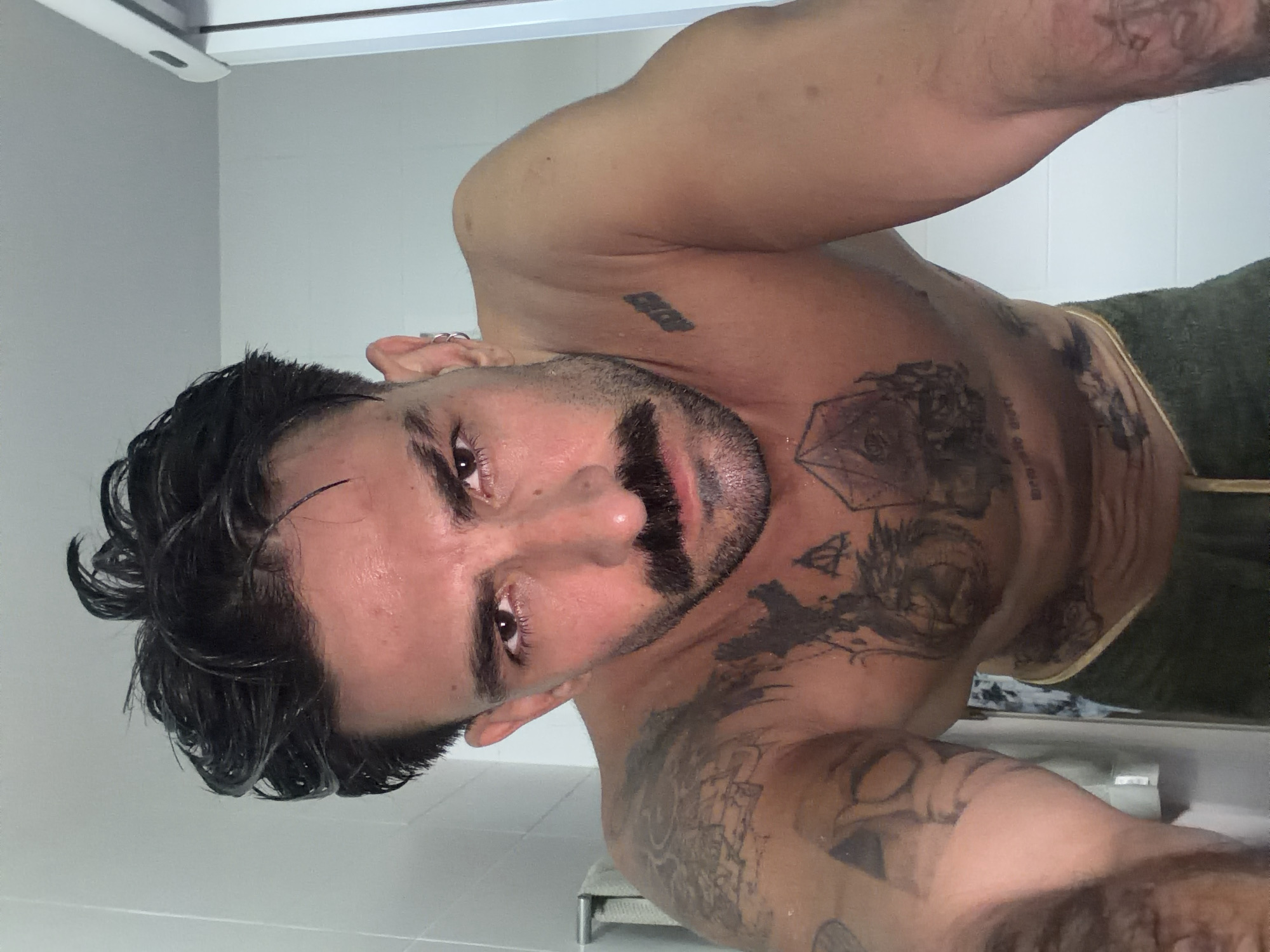 A man with dark, wet hair and a beard is taking a selfie in what appears to be a bathroom or shower. He is shirtless, revealing several tattoos on his chest and arms. He is wearing a dark-colored wrap around his waist. The background includes white tiles and a white light fixture. The picture was not taken in the city of Chalong, Thailand, so location is unknown.