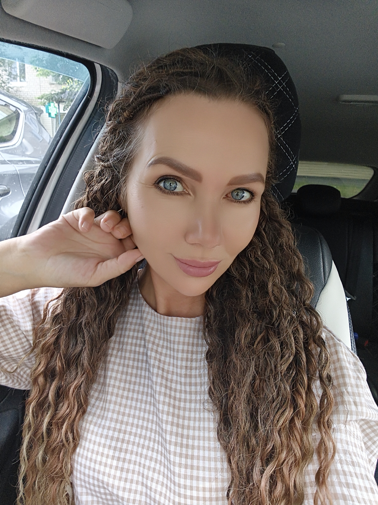 A close-up, indoor selfie shows a fair-skinned woman with long, curly brown hair styled into braids near her scalp, with the rest of her hair flowing down. Her right hand is raised to her face, gently touching her earlobe. She is wearing a beige and white checkered collared shirt. Her eyes, which are blue, are looking directly at the camera, and her lips are slightly parted, revealing a soft smile. The background shows the interior of a car, with a black and white car seat visible. The sun is shining outside, visible through the car window, and a building and trees can be seen in the distance. The overall impression is that the woman is in a car, possibly driving, and has taken a selfie.